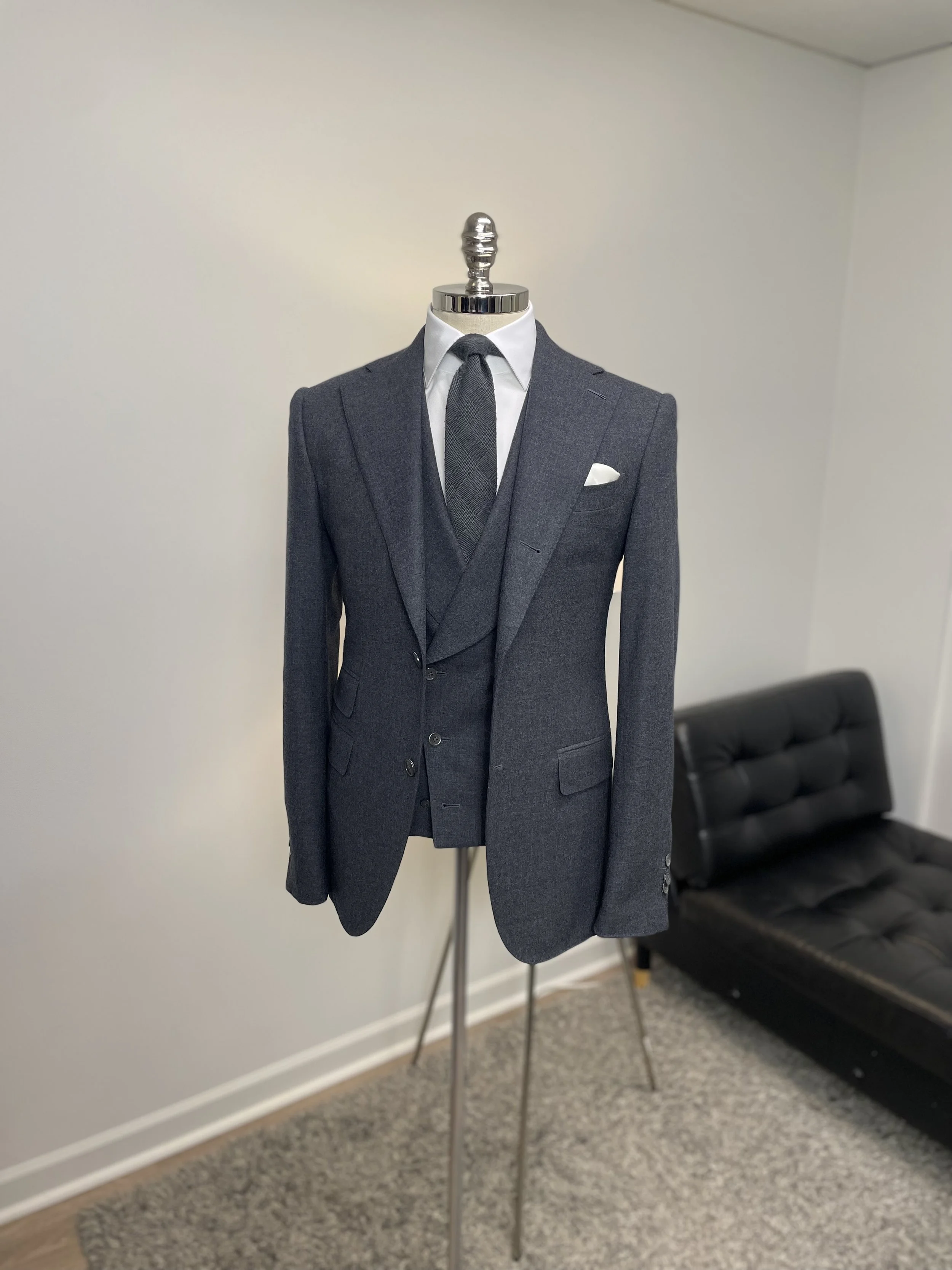 A mannequin dressed in a dark gray suit with a white dress shirt, a dark plaid tie, and a white pocket square, standing on metal legs in a room with a black sofa in the background.