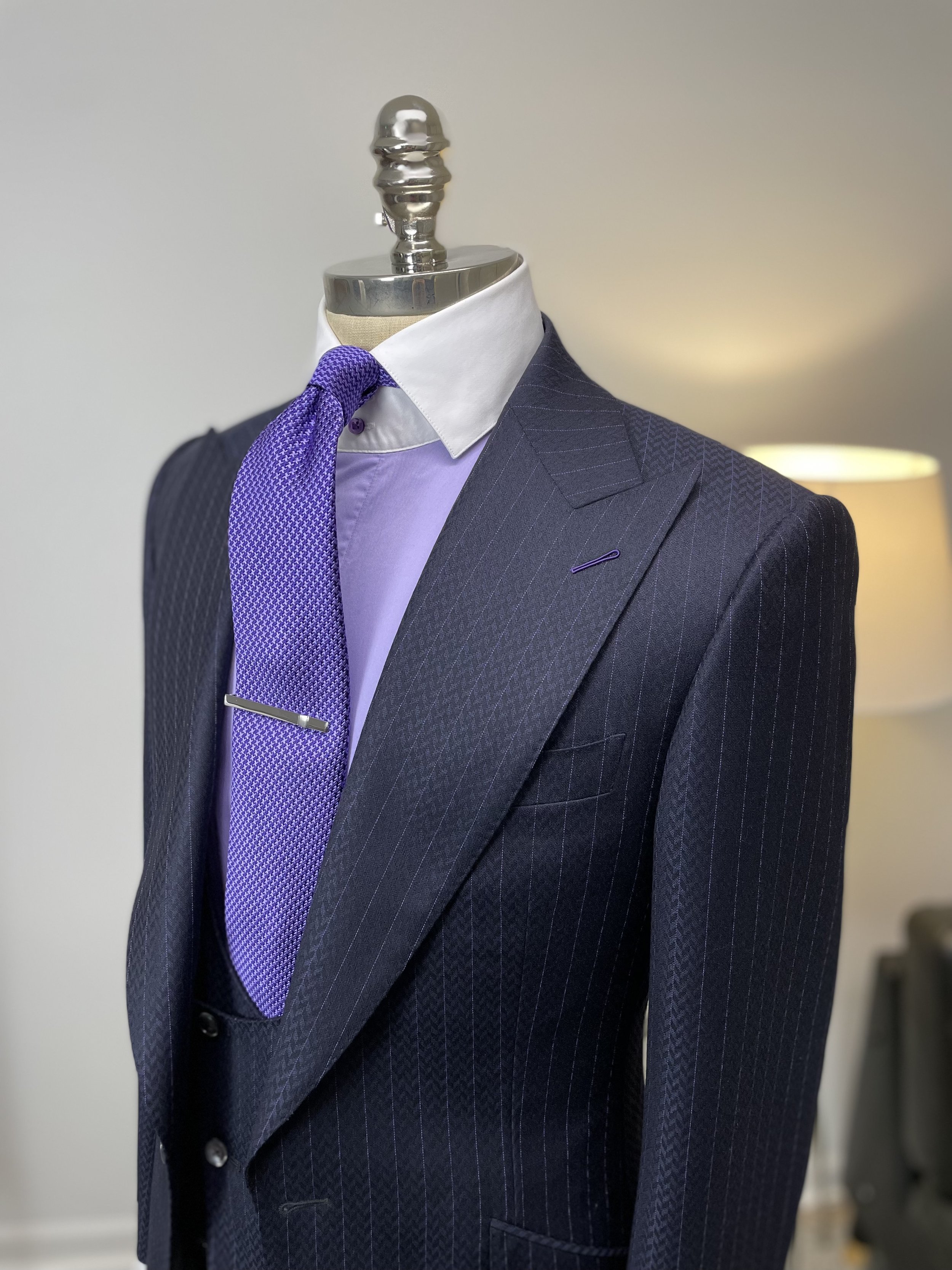 A mannequin dressed in a navy blue pinstripe suit with a white shirt, lavender tie with a silver clip, and a purple pocket square.