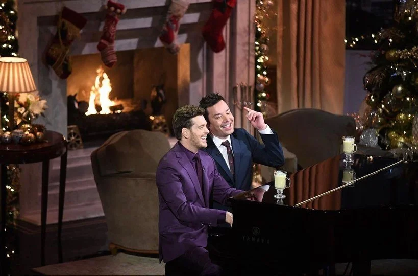 Two men (Michael Bublé and Jimmy Kimmel) in suits sitting at a black grand piano, smiling and enjoying music in a cozy, decorated living room with a fireplace and stockings hung above, Christmas tree, and festive lights.