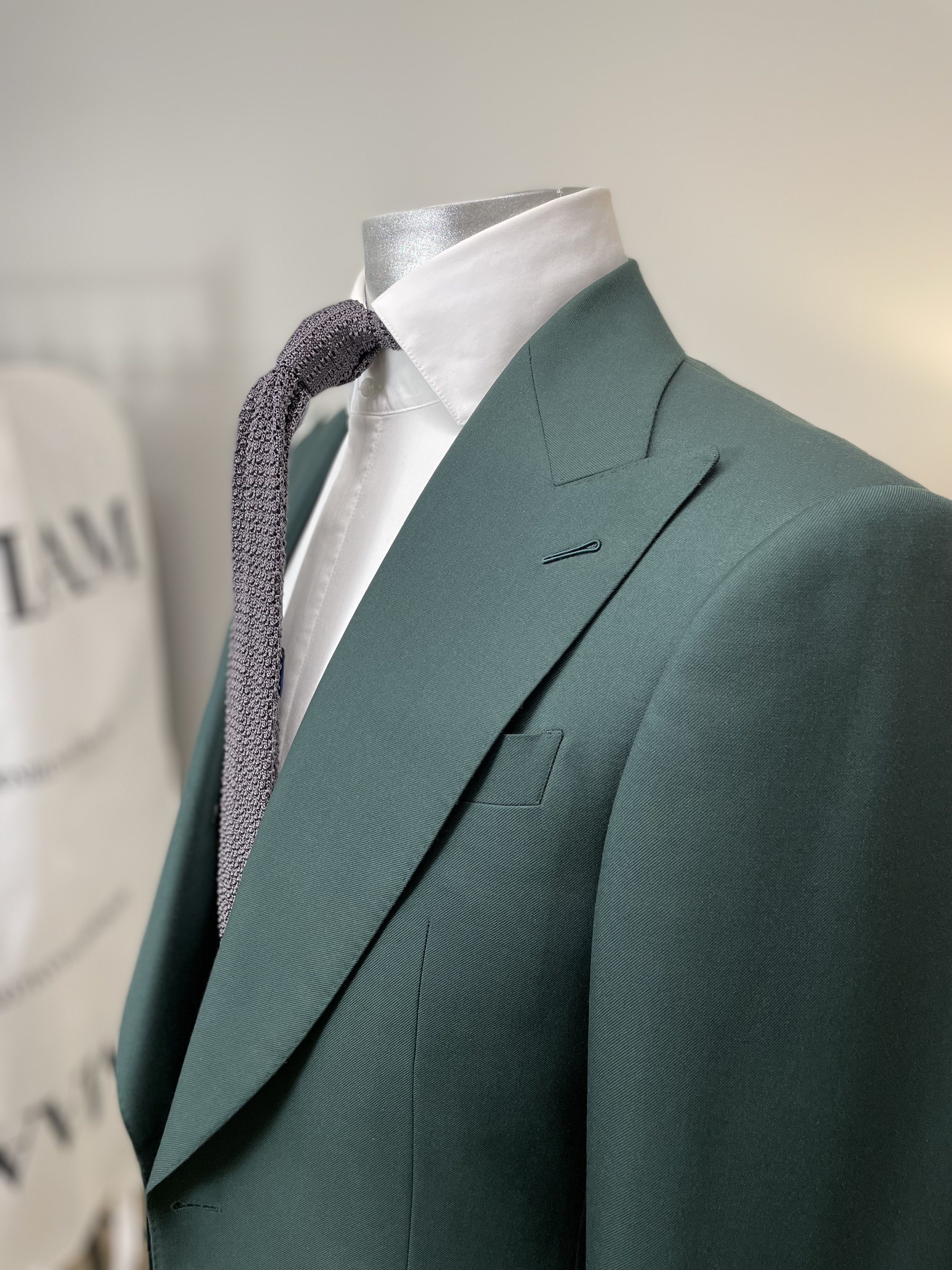 Mannequin dressed in a teal suit jacket, white shirt, and textured gray tie, with a blurred background showing part of a sign.