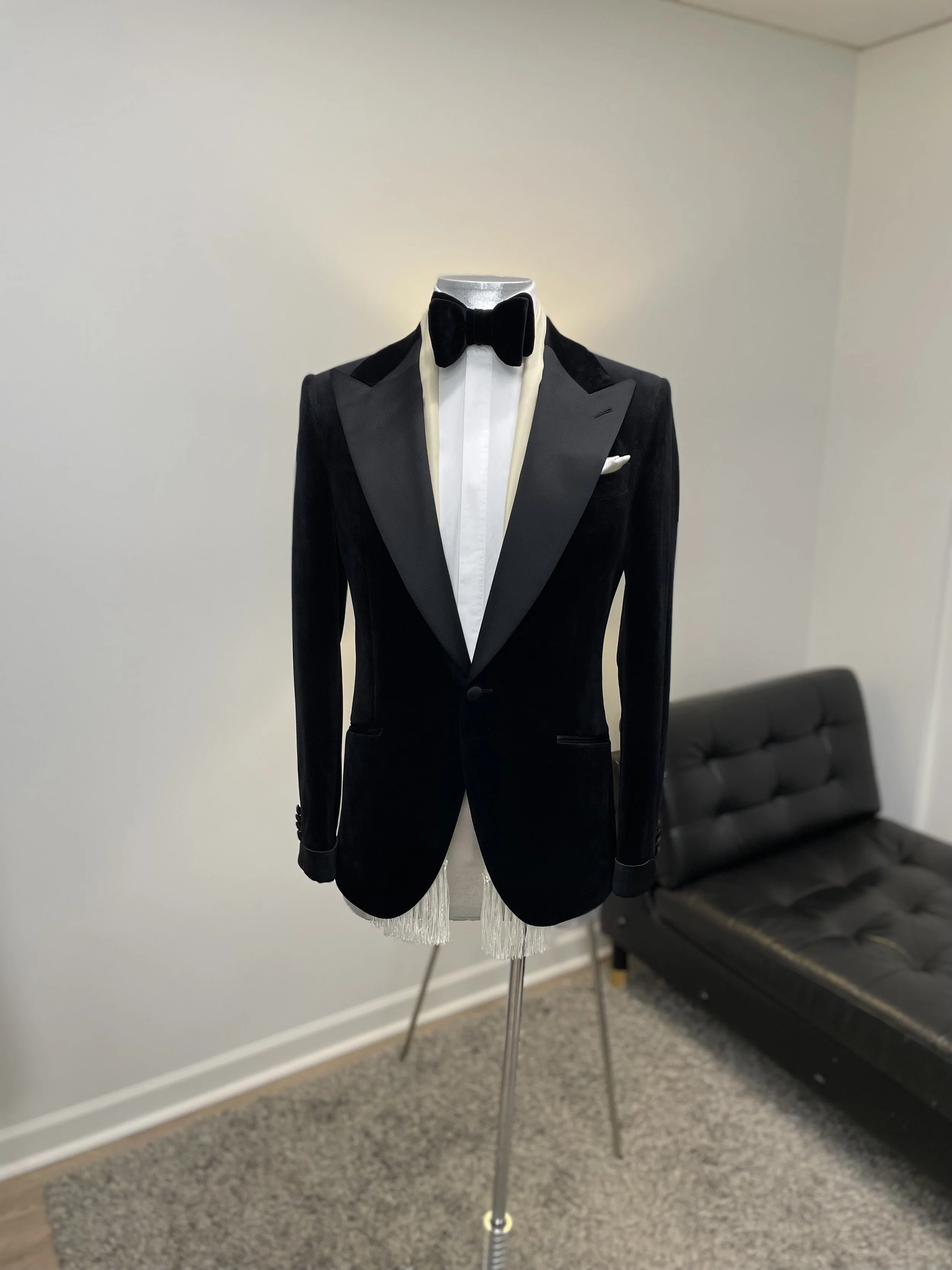 A mannequin dressed in a black tuxedo with a white shirt, black bow tie, and black velvet lapels, standing in a room with a gray wall and a black leather couch in the background.