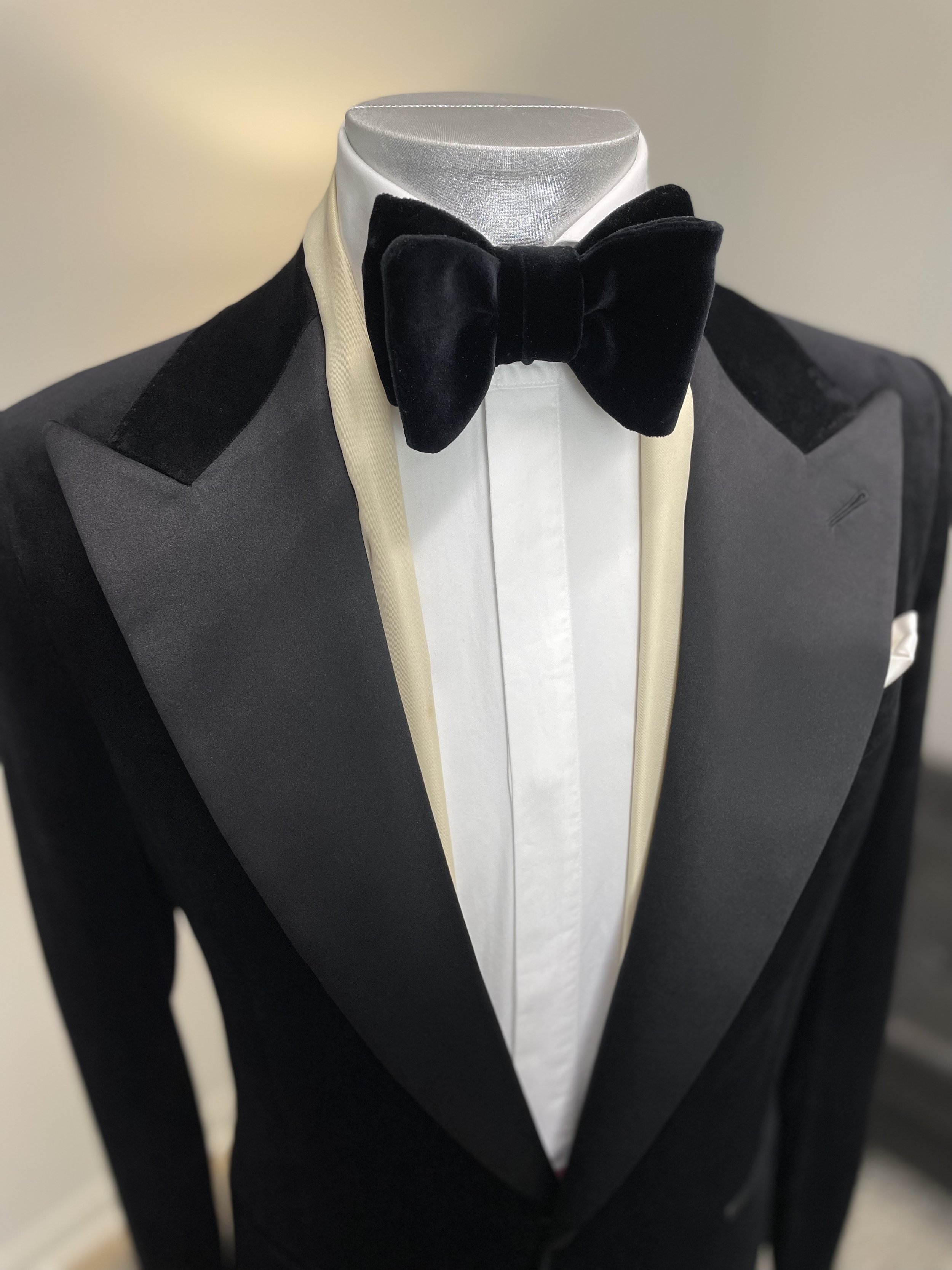 A tuxedo with black satin lapels, a white dress shirt, a cream-colored satin bib front, and a black velvet bow tie, displayed on a mannequin.