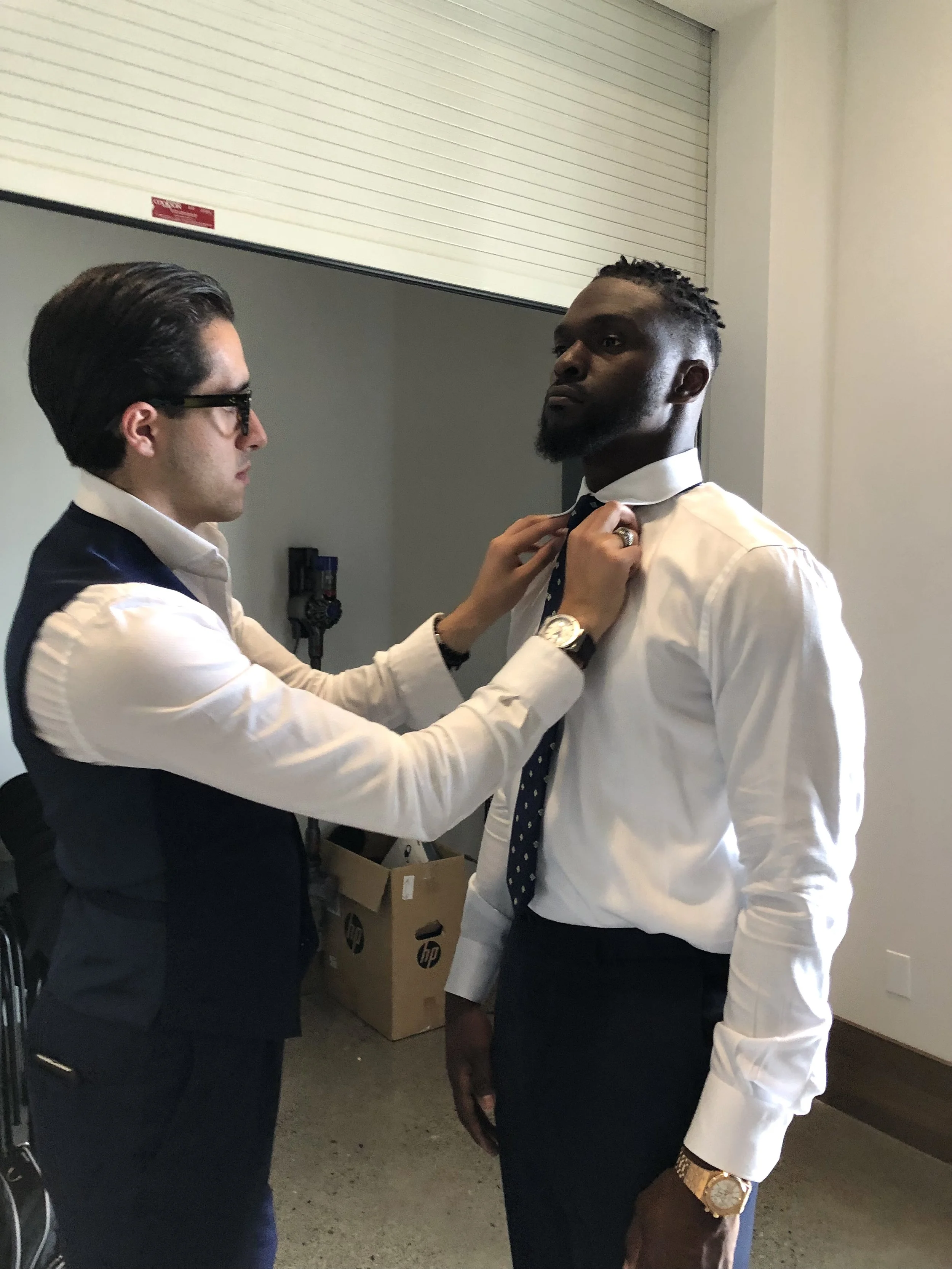 A man helps another man (Humaam and Cliff Avril) adjust his tie in a room. The man helping is wearing glasses and a vest with a white shirt, while the other man is wearing a white shirt, a navy tie with white dots, and a gold watch.