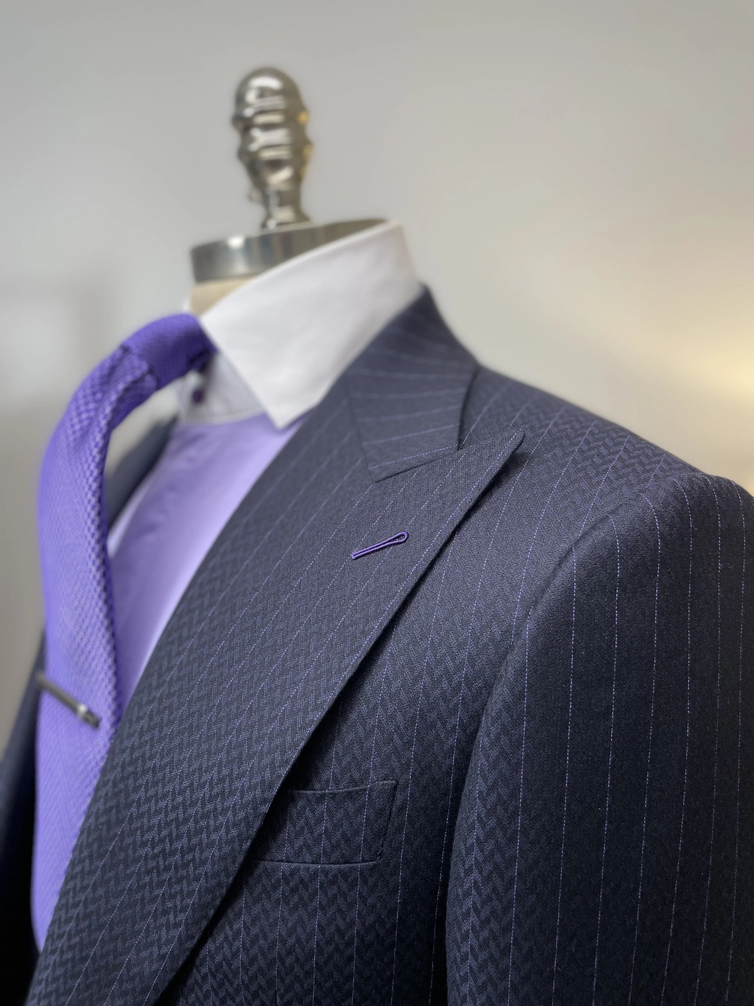 Close-up of a mannequin dressed in a dark gray pinstripe suit jacket, light purple shirt, white collar, and purple tie, against a neutral background.