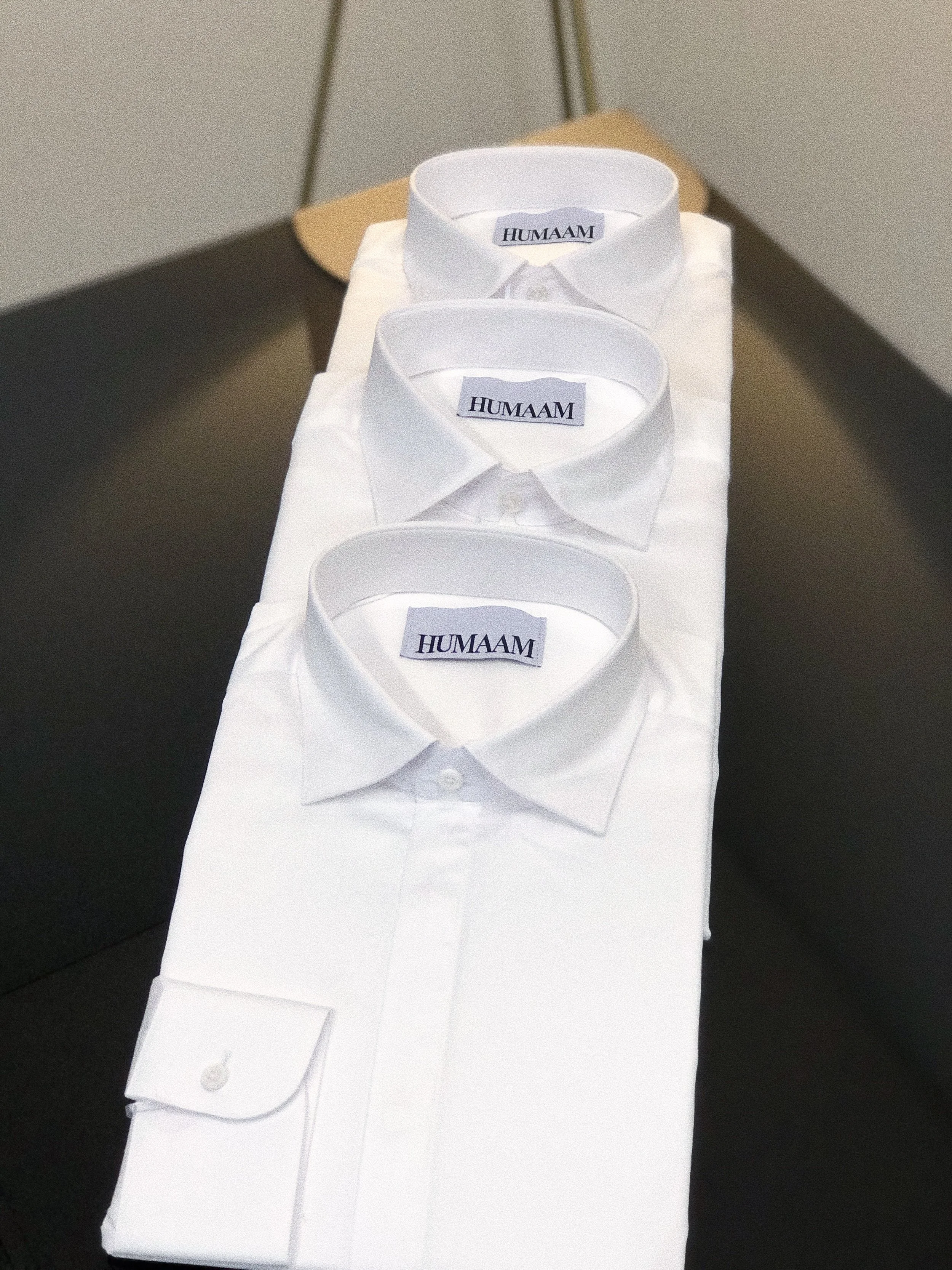 Three neatly folded white dress shirts with the brand label 'HUMAAM' visible on the collar, placed on top of a table or surface.