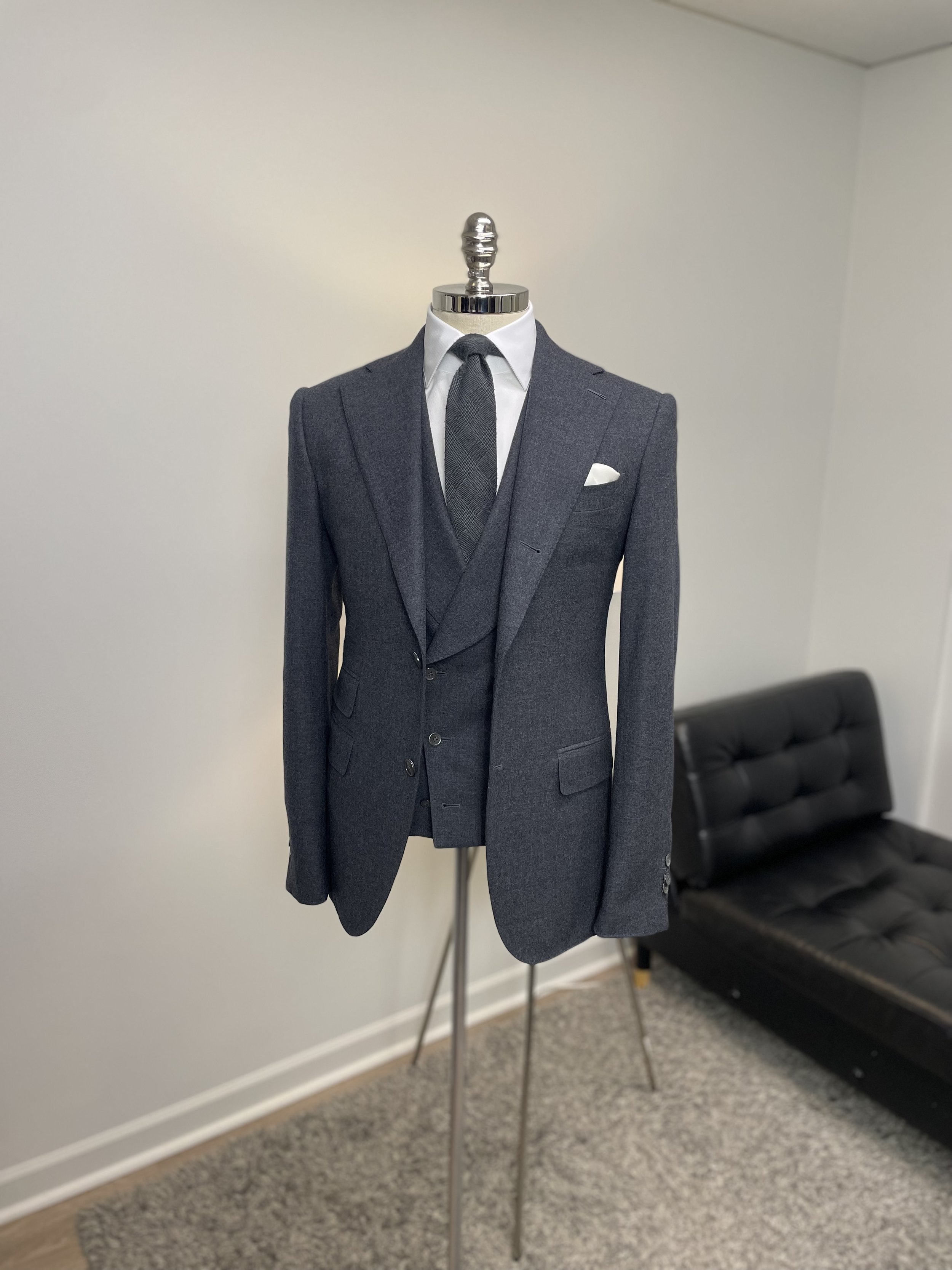 A mannequin dressed in a dark gray suit, with a white dress shirt, a dark gray tie, and a white pocket square, displayed on a stand in a room with a white wall and a black sofa in the background.