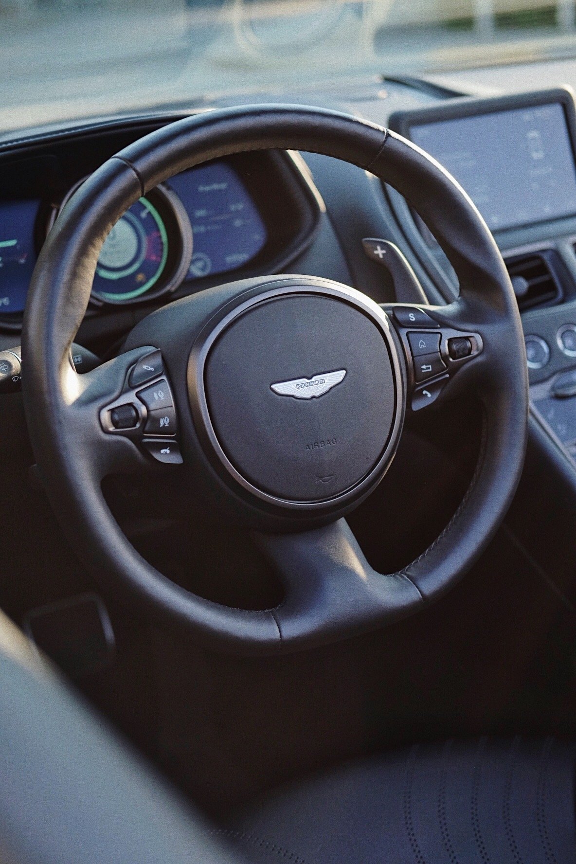 Close-up of an Aston Martin car steering wheel with digital dashboard and touchscreen display in the background.