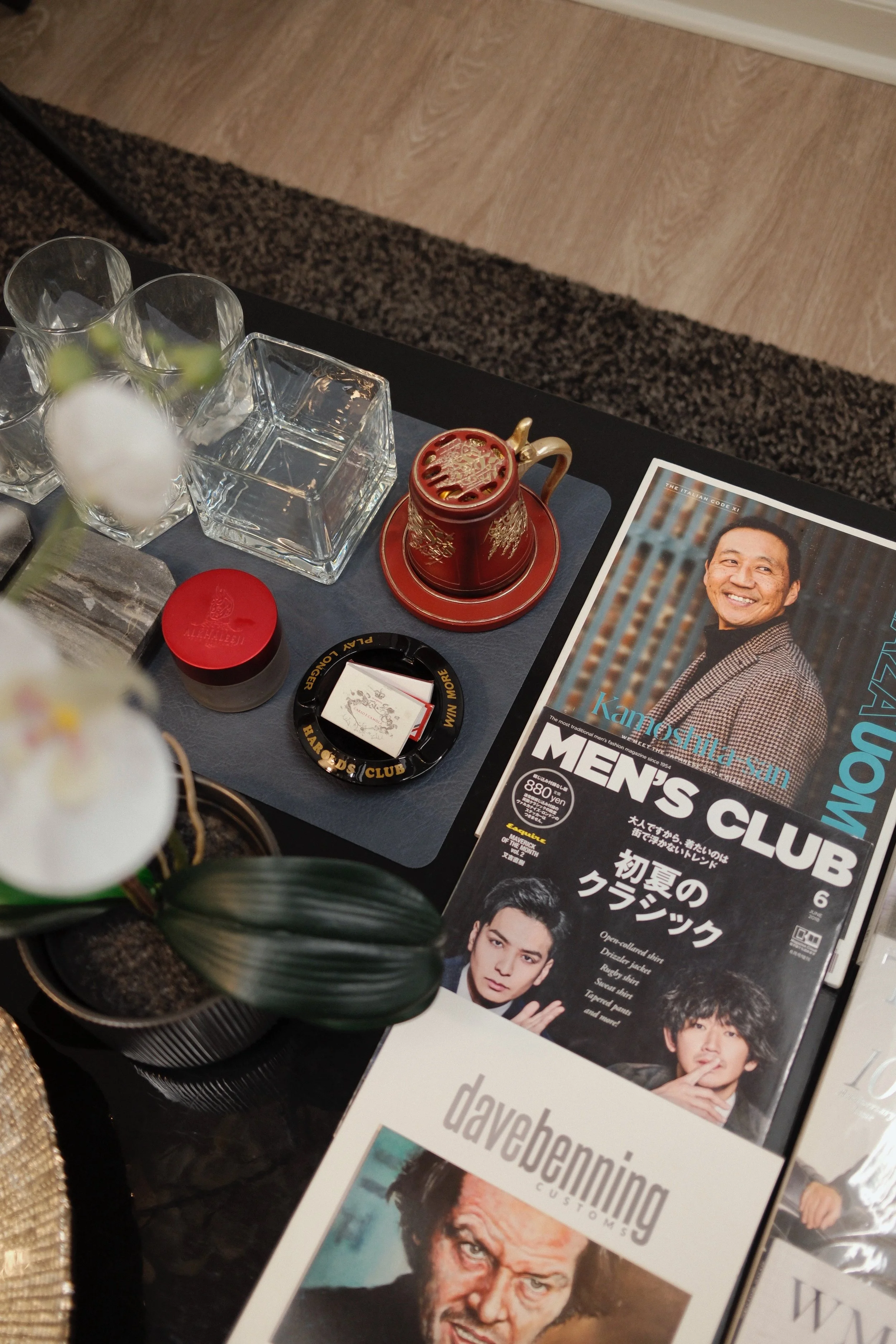 A table setup with clear glass cups, a red and gold decorative mug, a small red container, a black ashtray, and magazines featuring men's fashion and celebrity photos, with some floral decoration in the foreground.