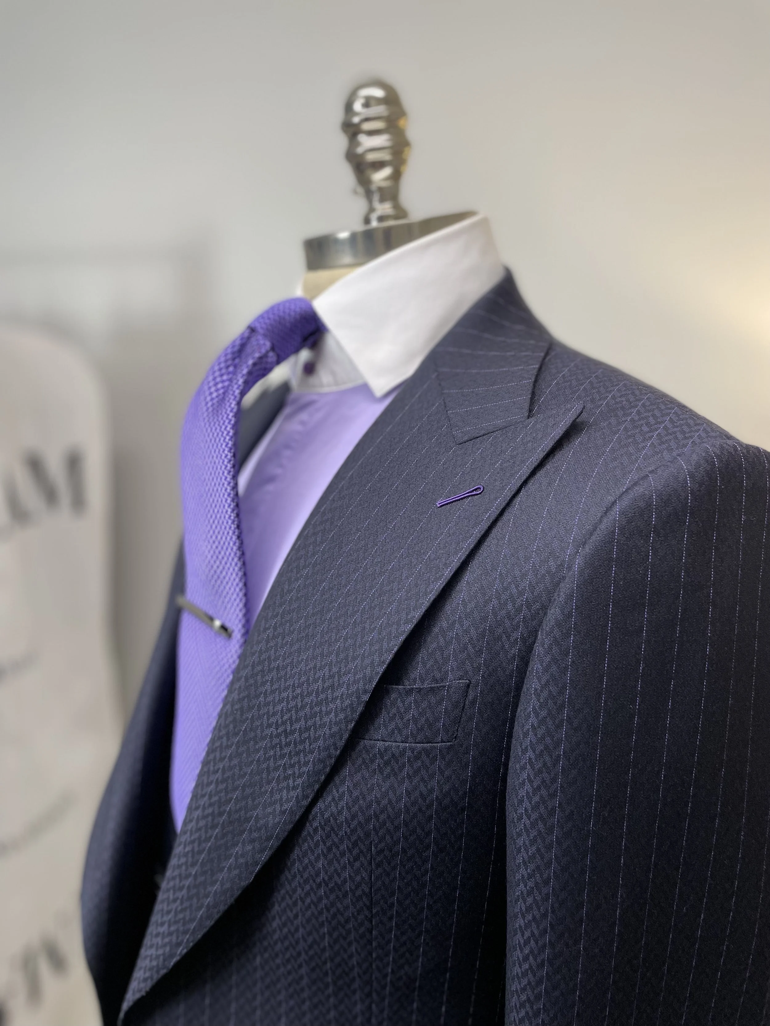 A mannequin dressed in a men's suit, white dress shirt, purple tie, and a purple pocket square, displayed on a mannequin with a metallic neck, against a plain light-colored background.
