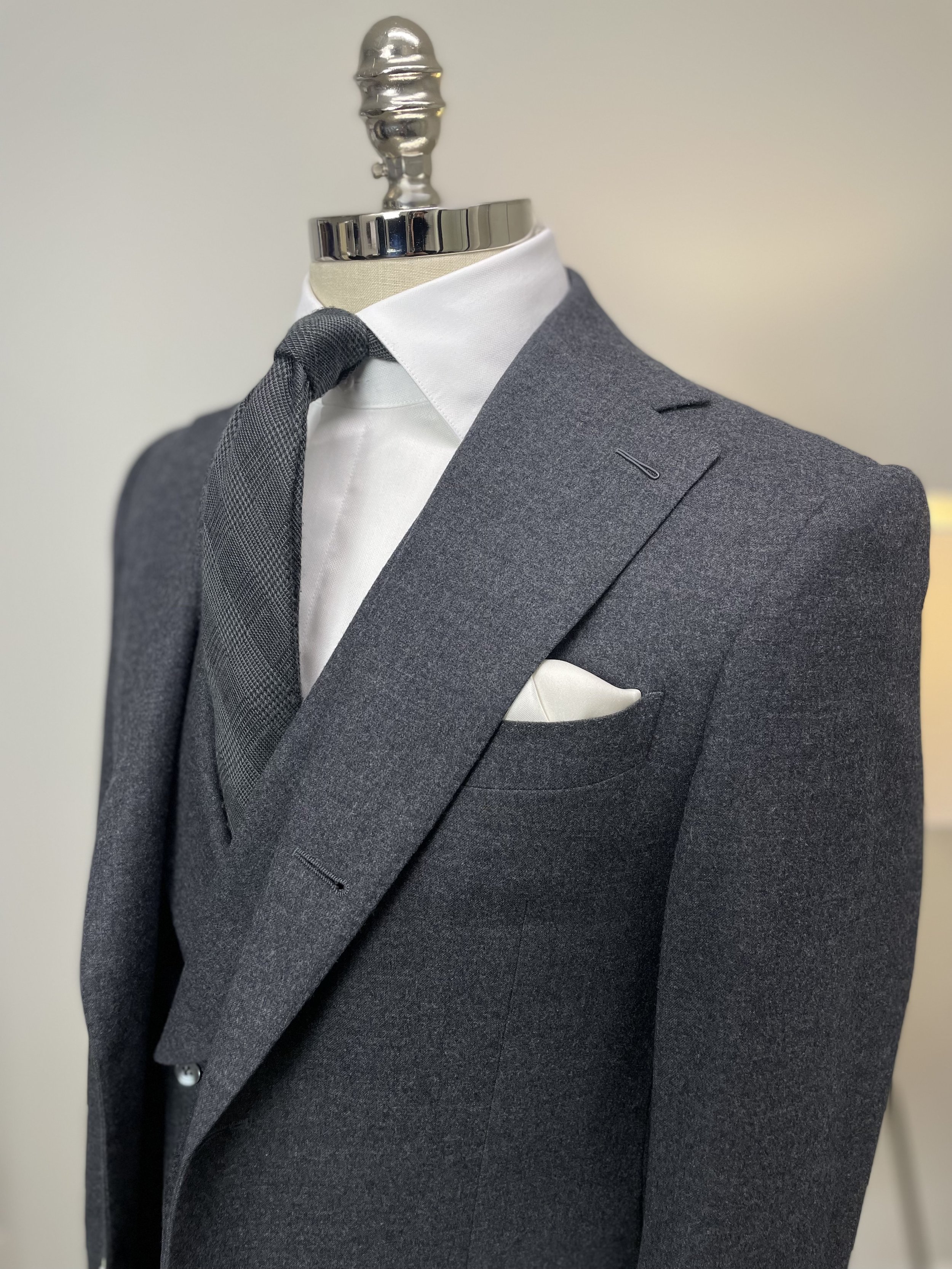 Mannequin dressed in a gray suit with a white dress shirt, a dark gray tie, and a white pocket square. The mannequin has a metallic head and neck, displayed against a plain light-colored background.