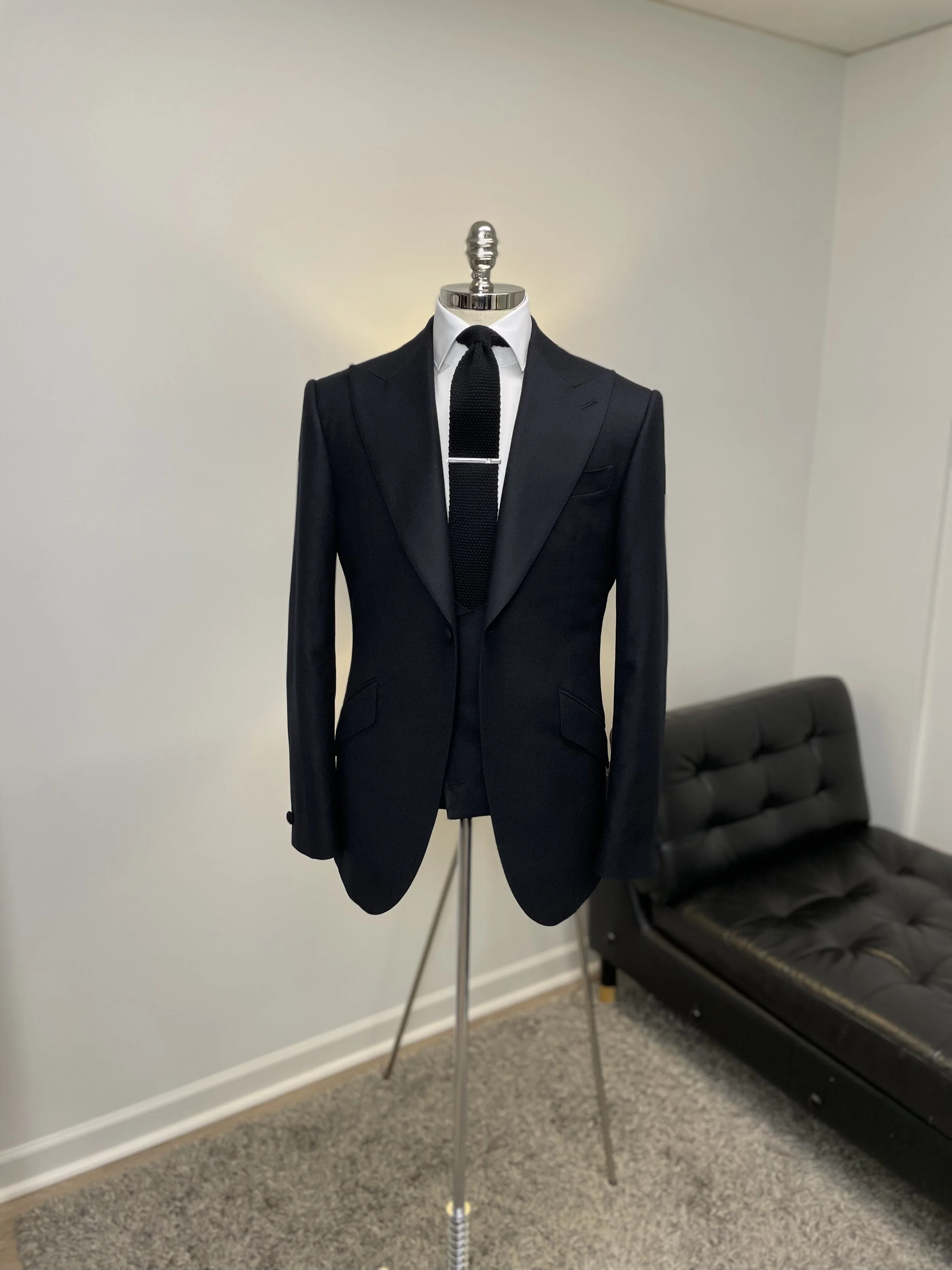 A mannequin dressed in a black suit with a white shirt and black tie, placed in a room with beige carpet and a black leather couch in the background.