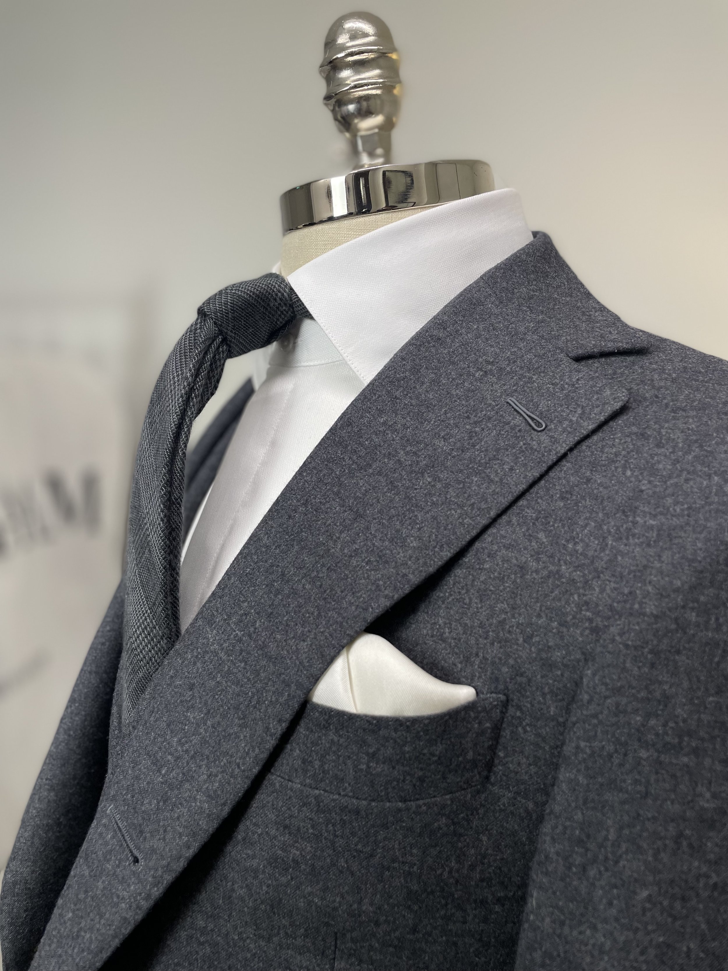 Close-up of a men's dark gray suit jacket with white dress shirt, gray tie, and white pocket square, displayed on a mannequin.