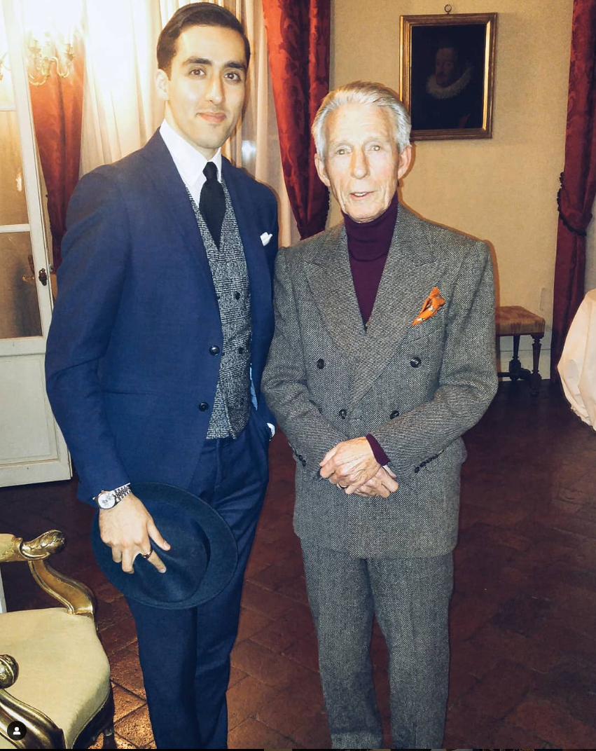 Two men side by side in a room with elegant decor, one younger (Humaam) and one older (Edward Sexton), both dressed in formal attire, with a painting on the wall behind them.