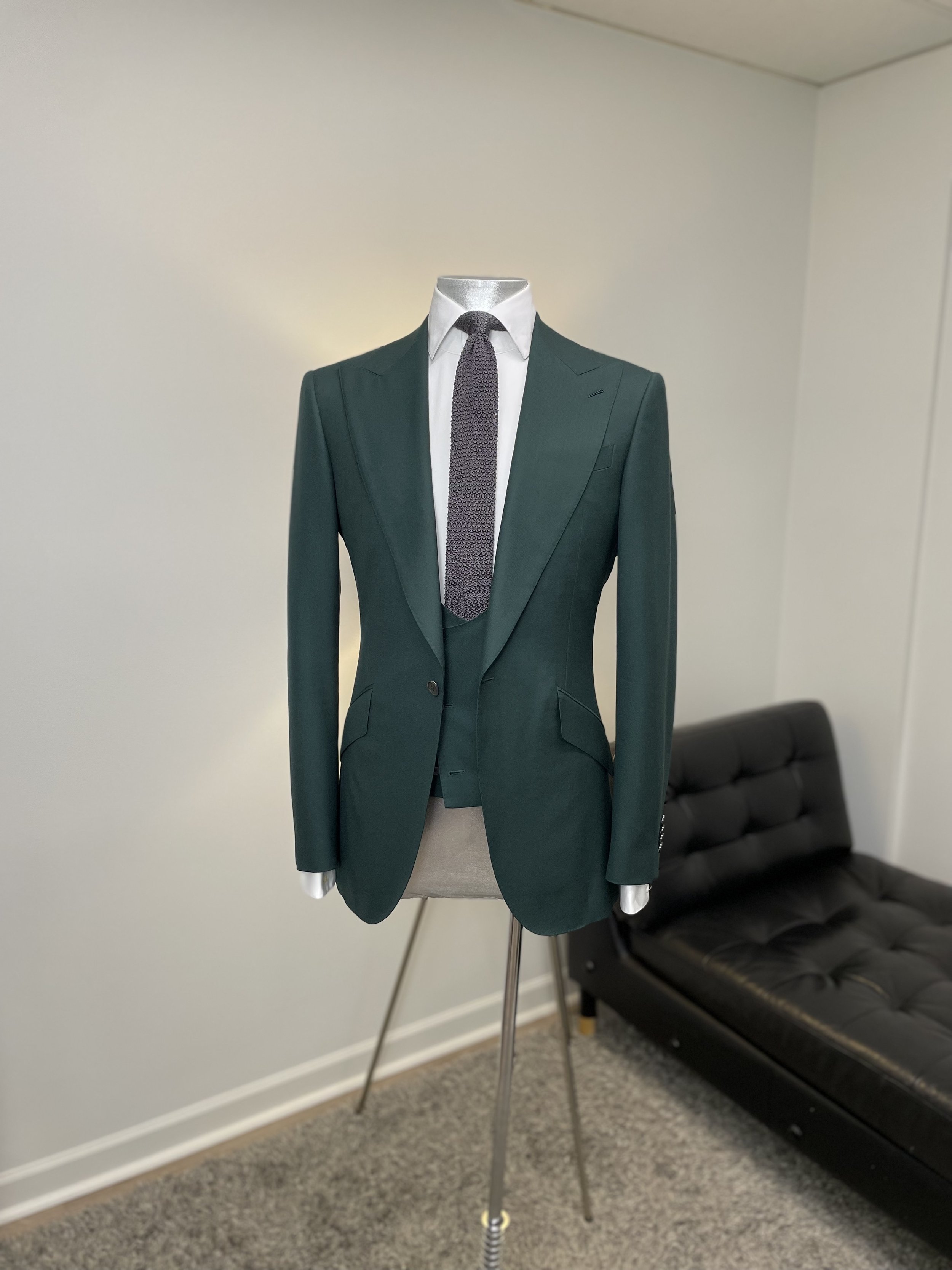 A mannequin dressed in a dark green blazer with a white shirt and a grey tie, standing in a room with a black couch and a plain white wall.