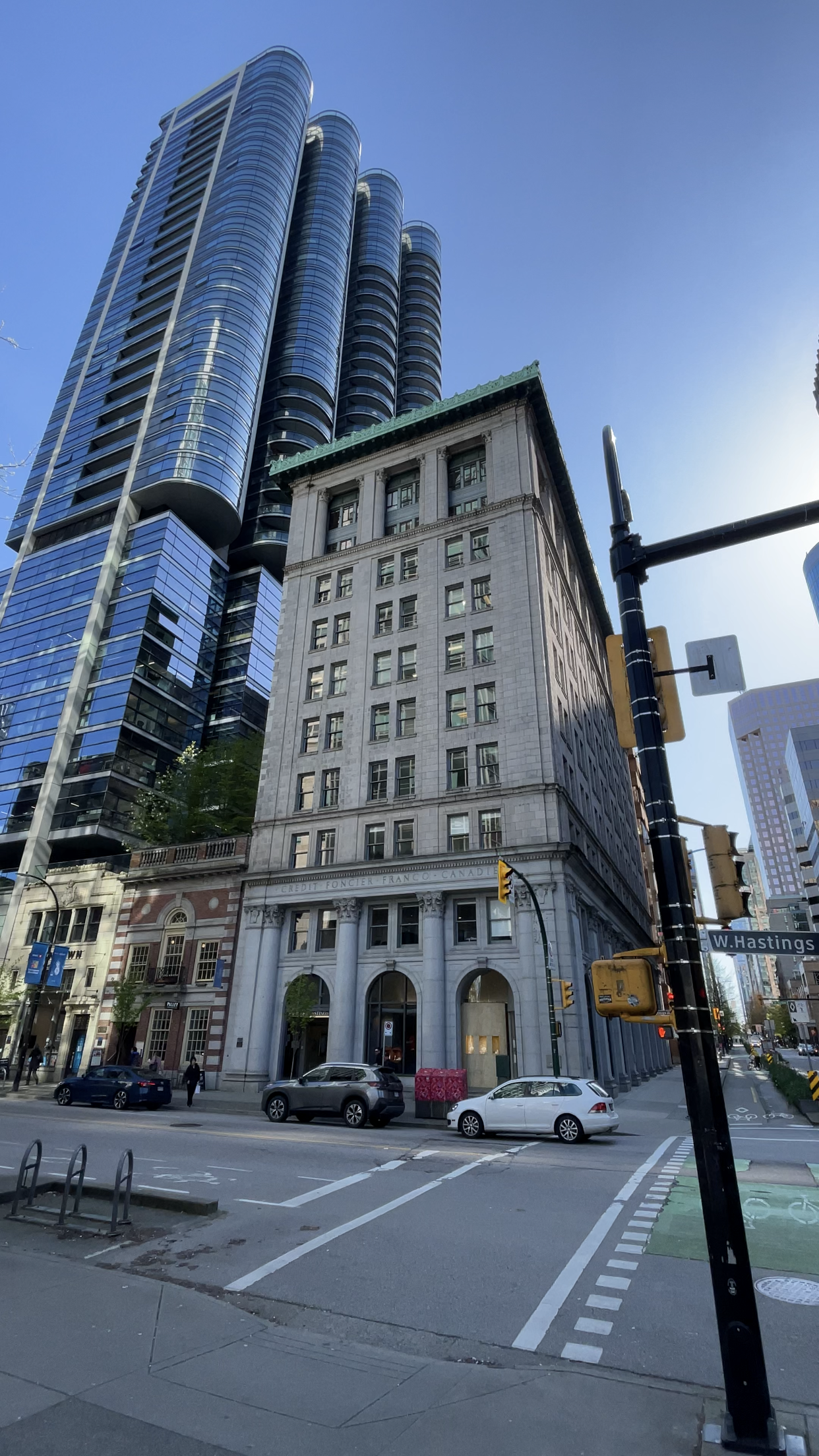 Street view of a city intersection with tall modern skyscrapers and a historic building, cars parked along the street, and pedestrians walking.