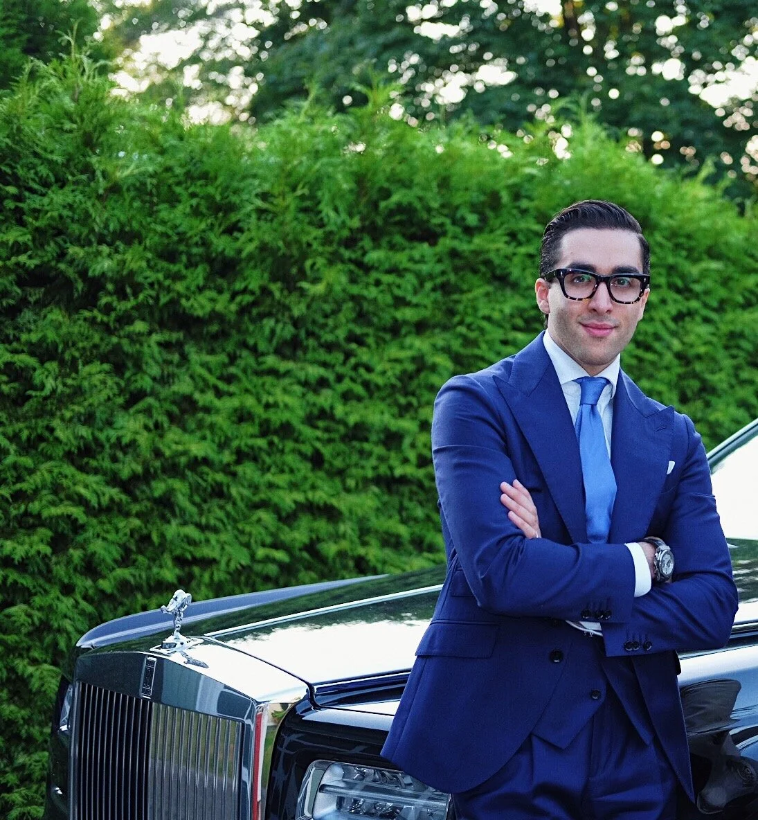 A man in a blue suit and glasses standing in front of a black luxury car, with arms crossed, outdoors with green bushes and trees in the background.
