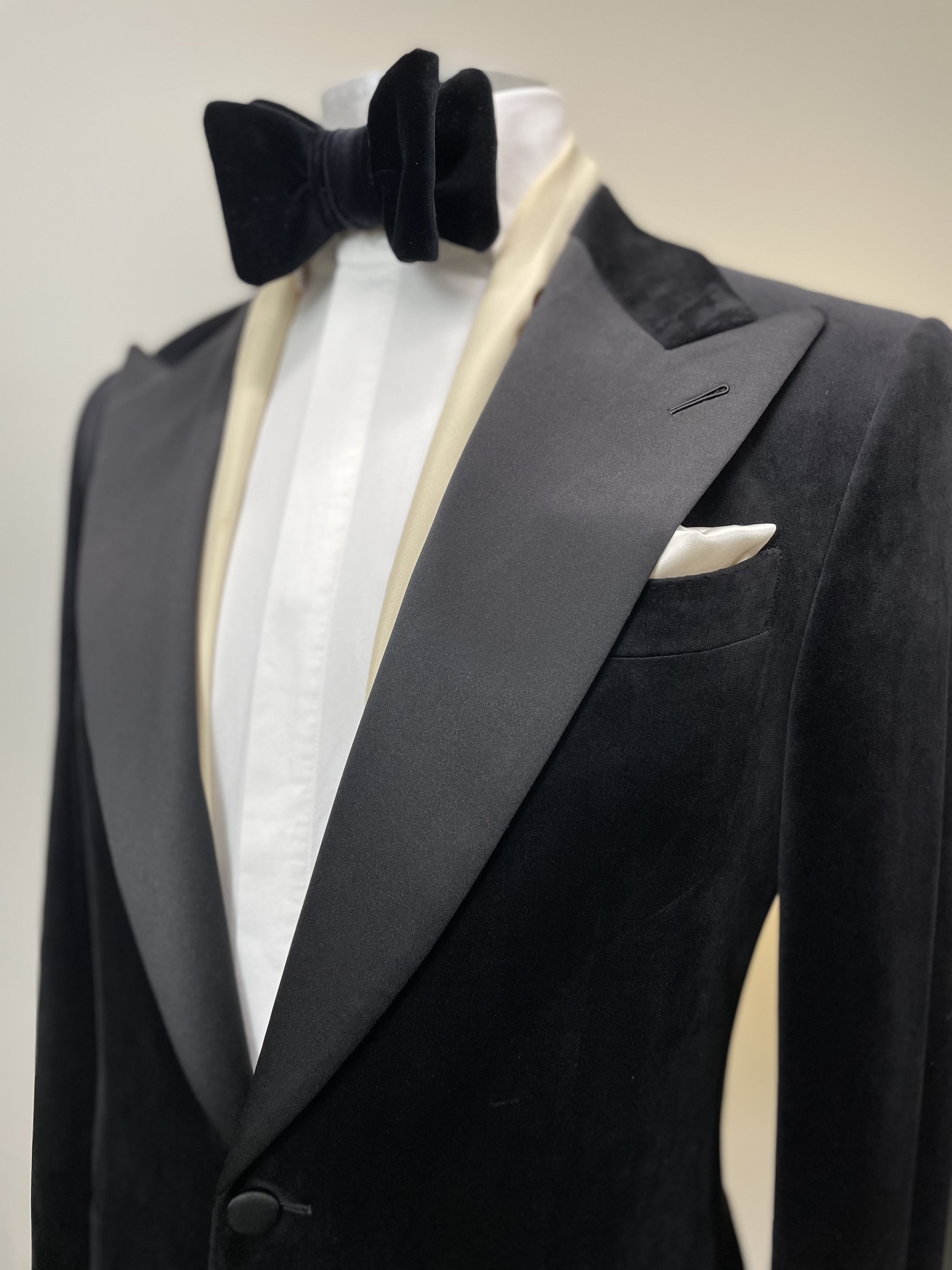 Black tuxedo jacket with satin lapel, white dress shirt, black velvet bow tie, and a white pocket square on a mannequin.