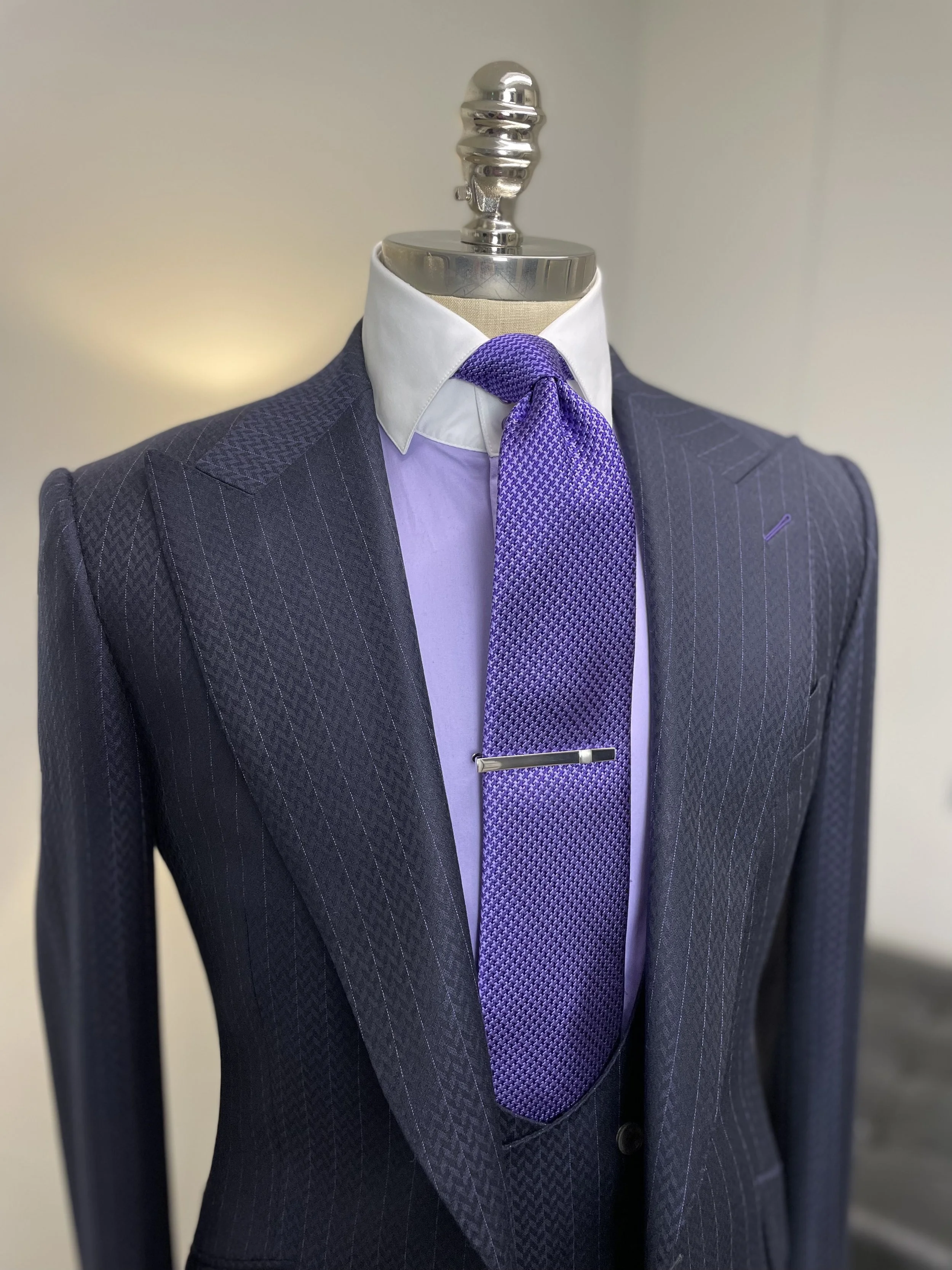 A mannequin dressed in a dark pinstripe suit, white shirt, purple tie with a tie clip, and a white collar, displayed in a retail store or showroom.