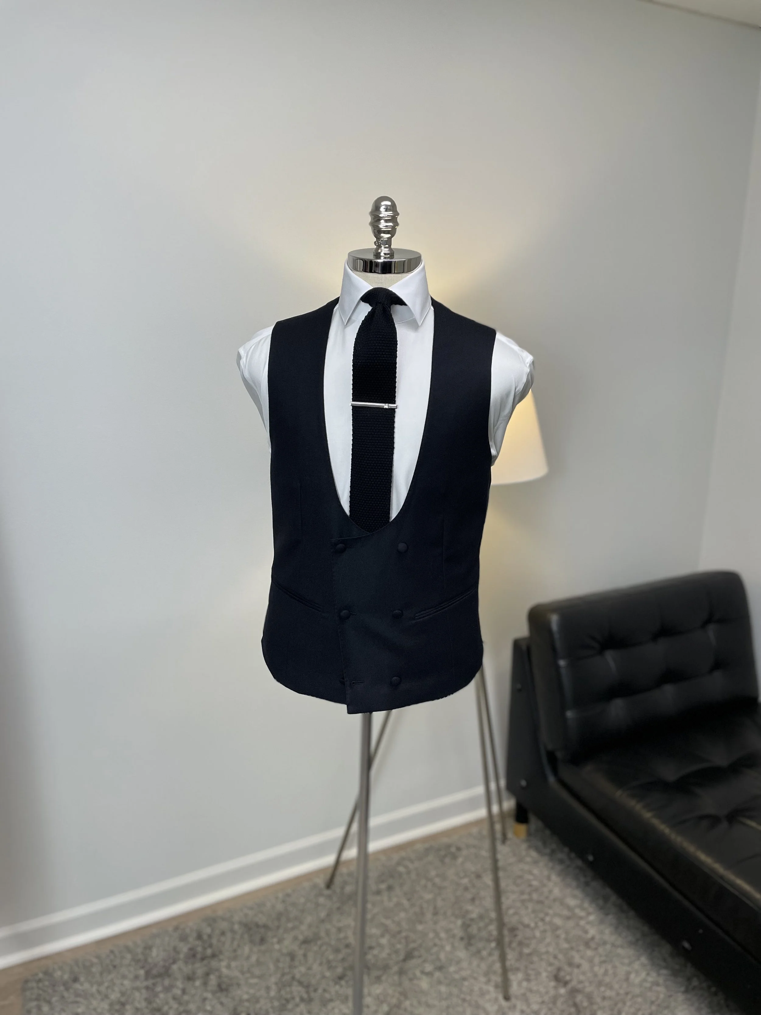 A mannequin dressed in a white shirt, black vest, and black tie, standing indoors with a lamp and a black sofa in the background.
