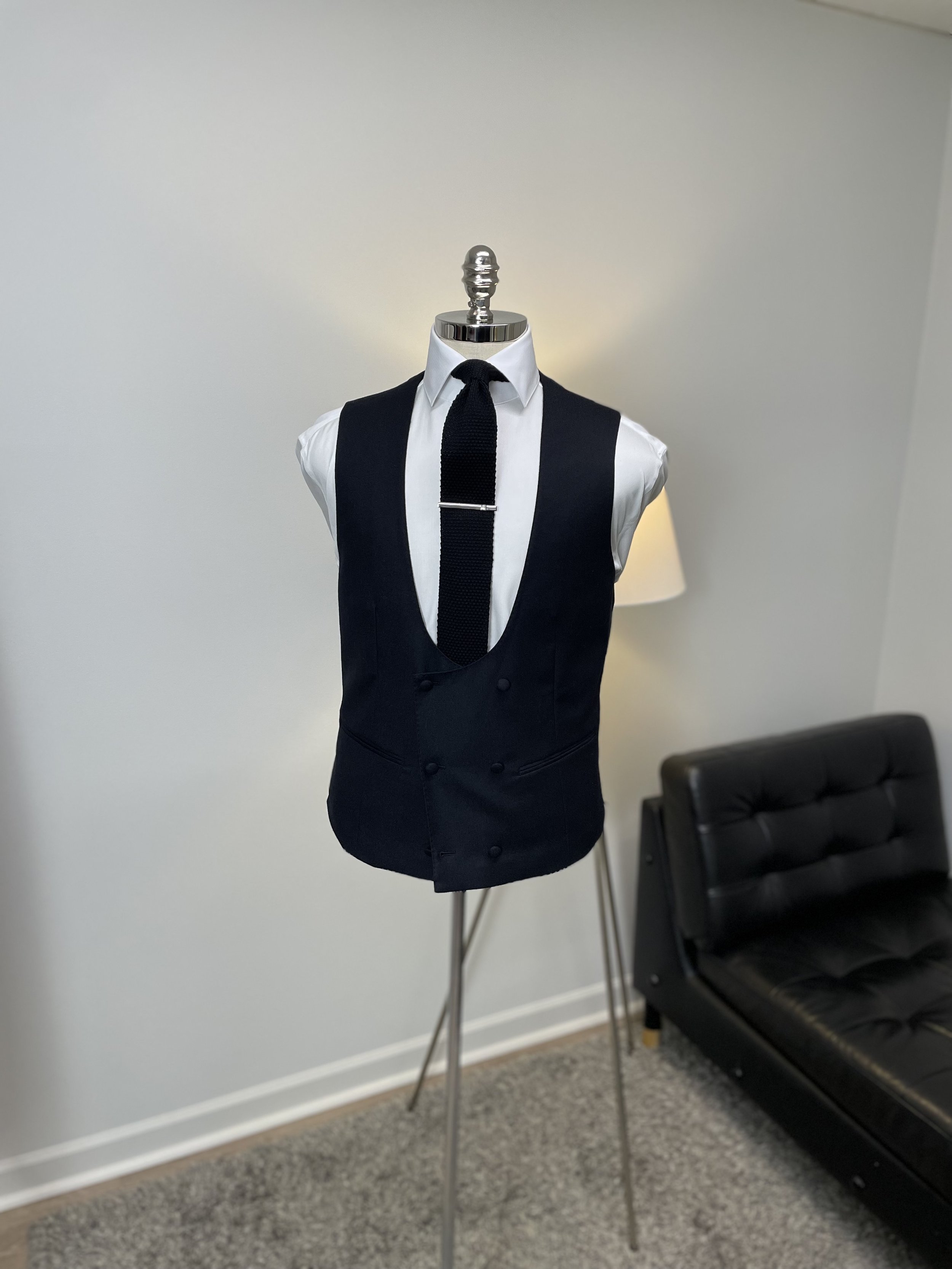 A mannequin dressed in a black tuxedo vest, white dress shirt, and black tie, with a tie clip, standing in a room with a black couch and a standing lamp.