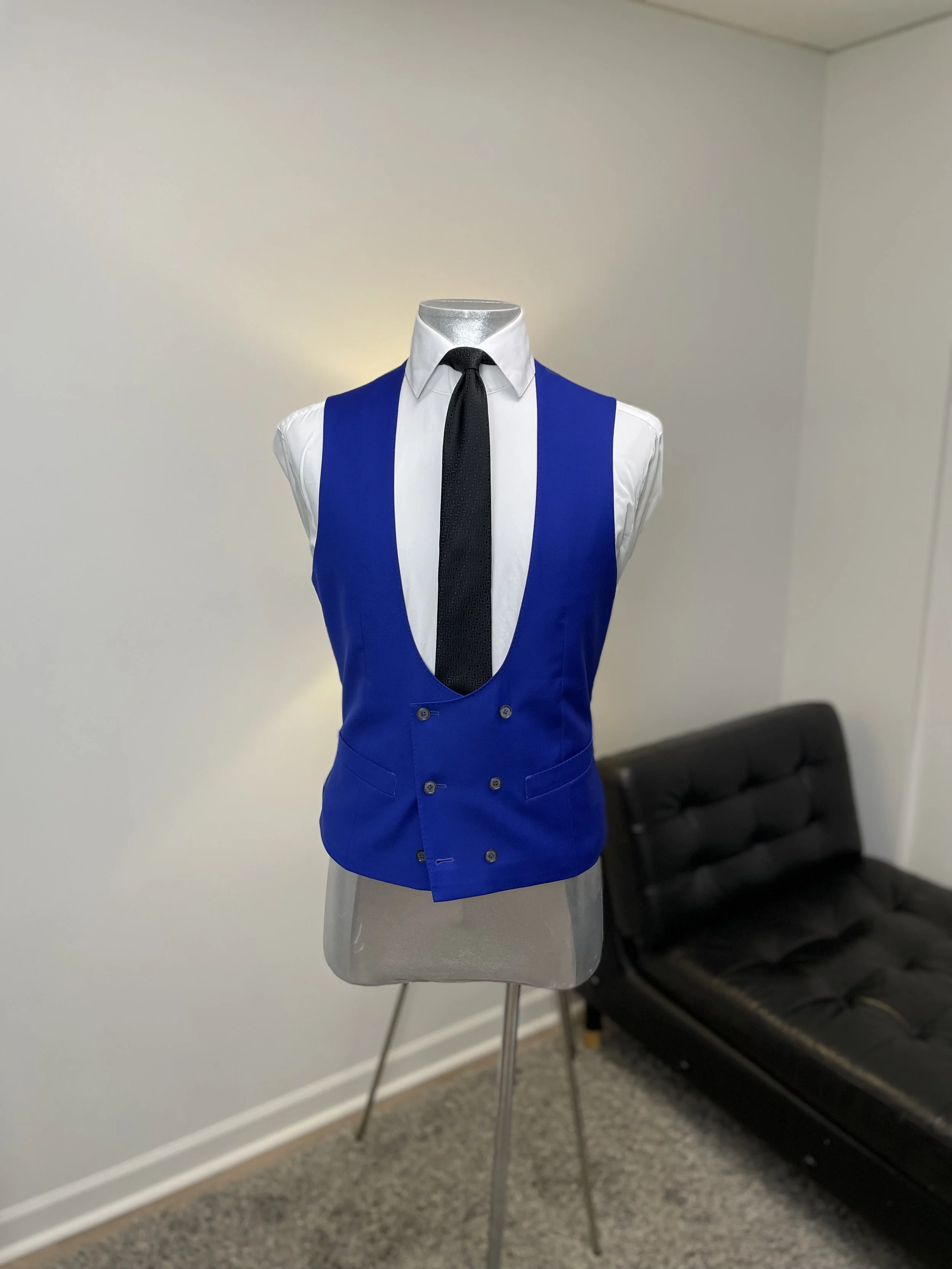 Mannequin dressed in a white shirt, blue vest, with a black tie, in a room with beige carpet, white walls, and a black leather couch.