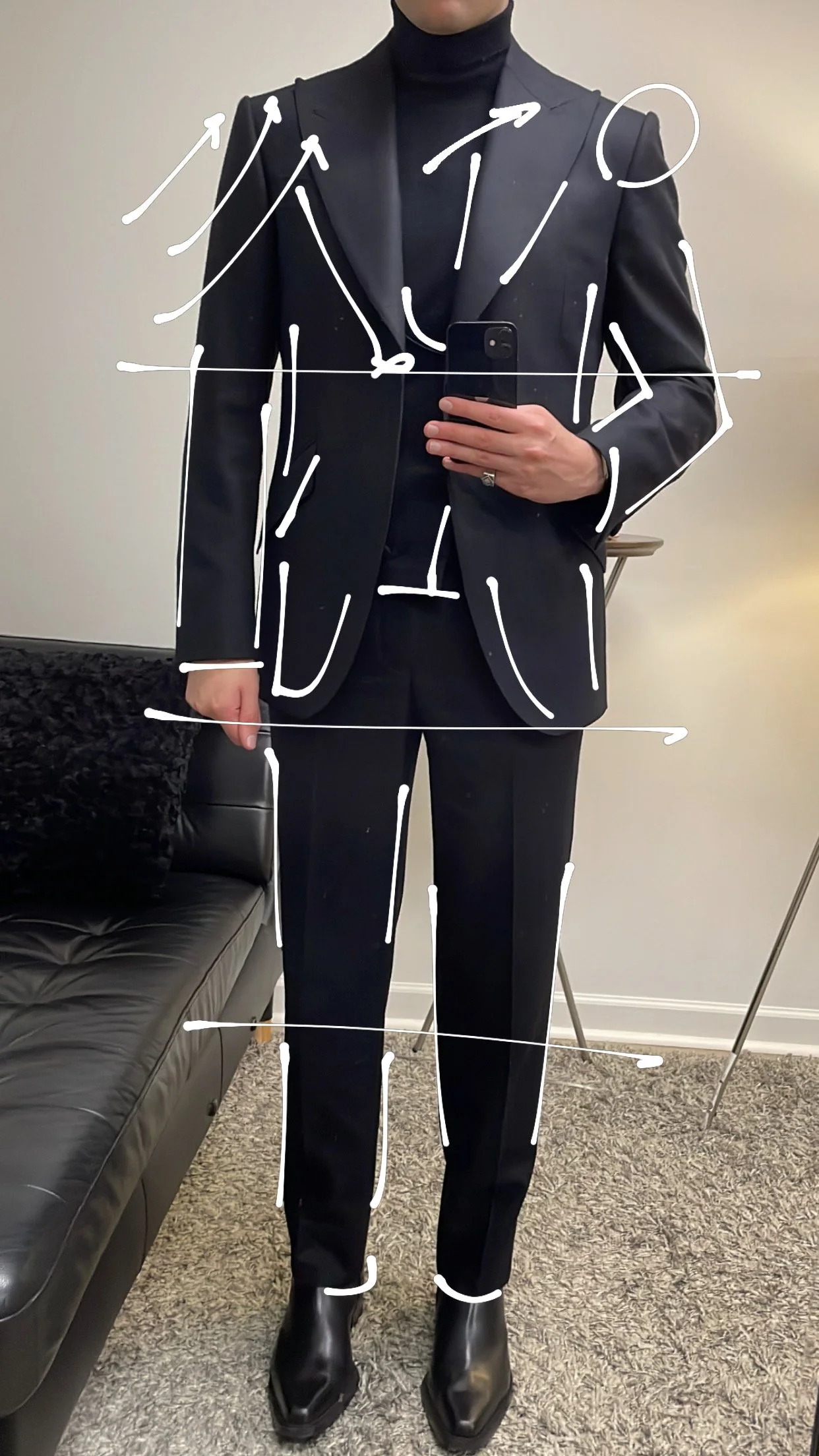 A person taking a mirror selfie in a black suit with white chalk markings and arrows pointing to different parts of the outfit, wearing a black turtleneck, black pants, and black boots.