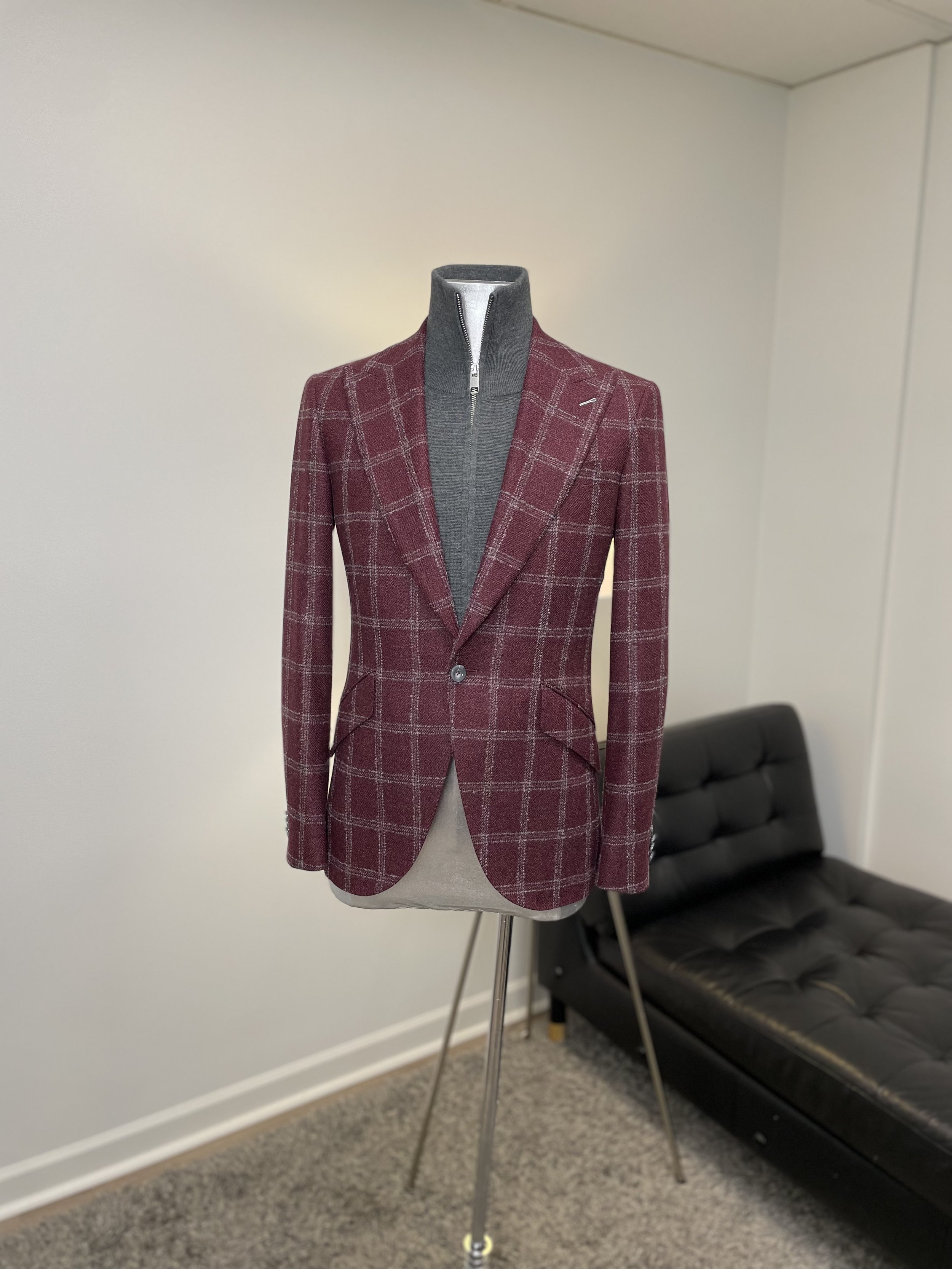 Men's blazer with a red and gray plaid pattern on a mannequin, layered over a gray zip-up sweater, standing in a room with a gray wall and black couch.