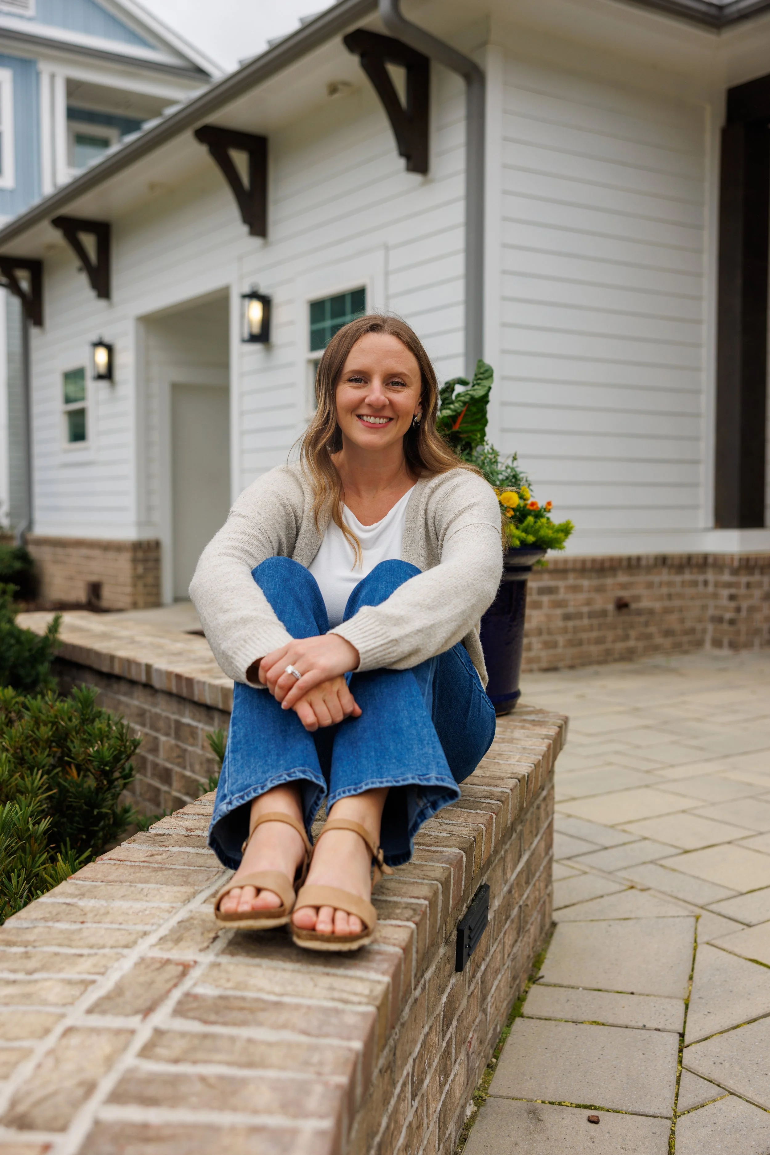 woman smiling with light brown hair sitting on a short brick wall with her arms wrapped around her legs