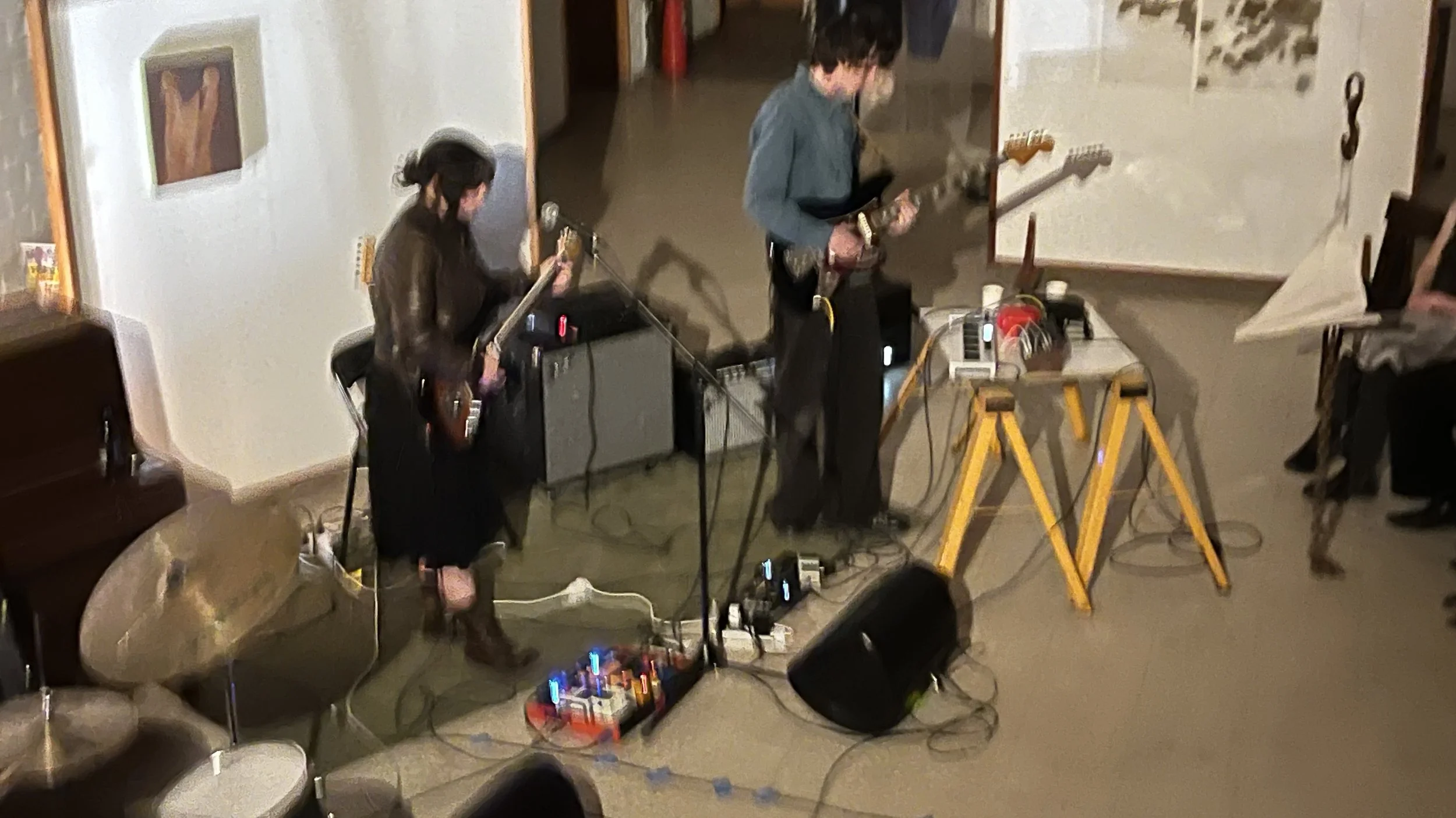 Two musicians, a woman and a man, performing with electric guitars in an indoor setting, with musical equipment and a whiteboard in the background.