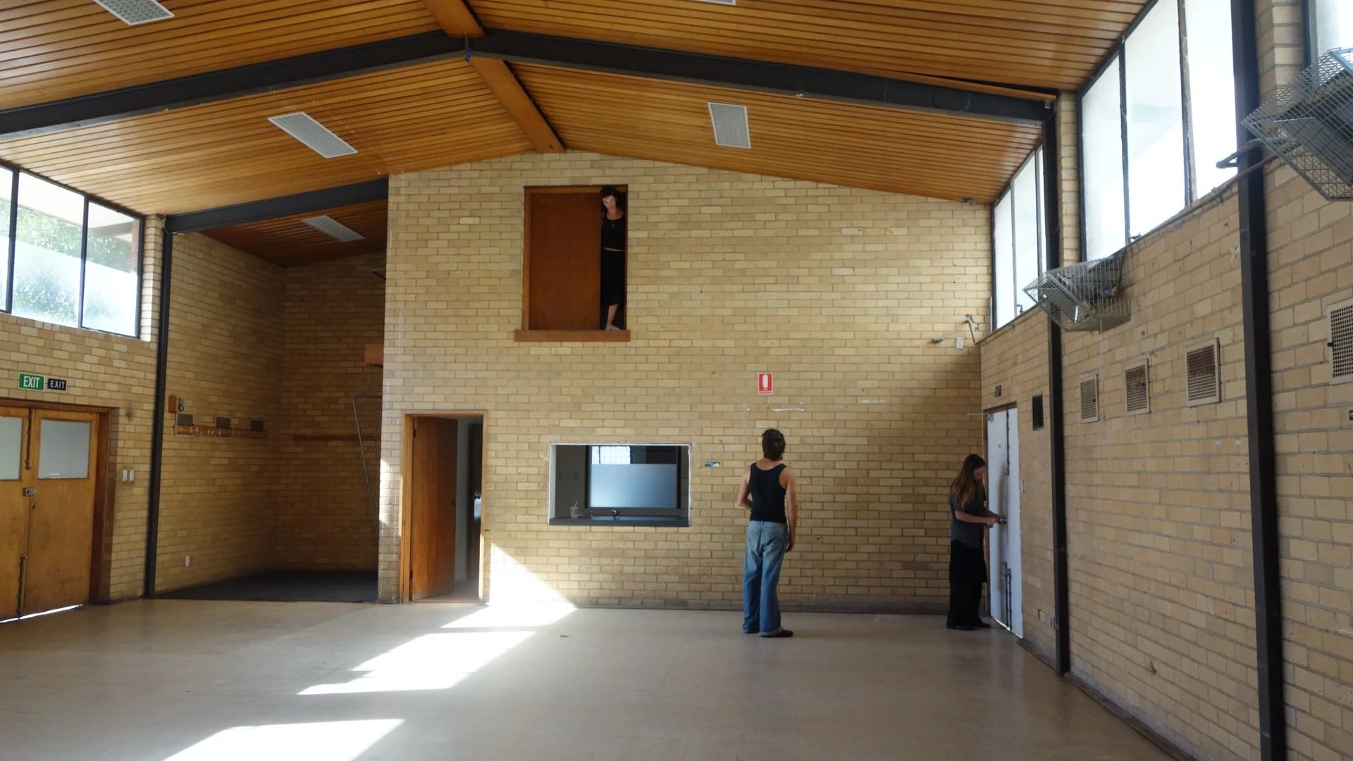 Interior of a spacious room with brick walls, wooden ceiling, and large windows; three people are present, one woman looking out from a second story window, one man standing and looking up, and one woman near a door.