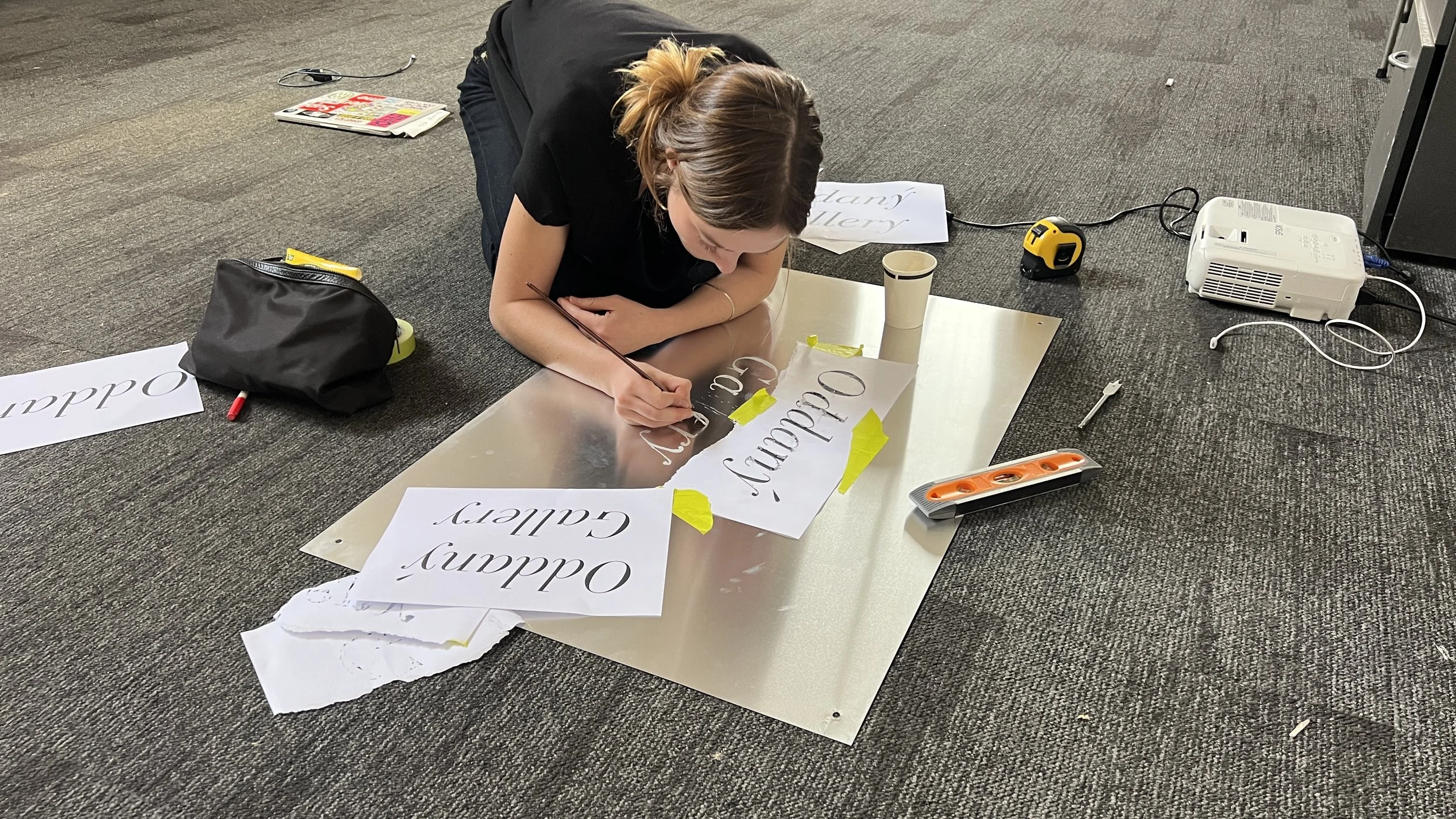 A woman is lying on the floor lettering a sign with white paper that says Gallery" in black calligraphy. There are additional sheets with the same text, a black bag, a cup, a tape measure, an extension cord, a projector, and a power strip around her.