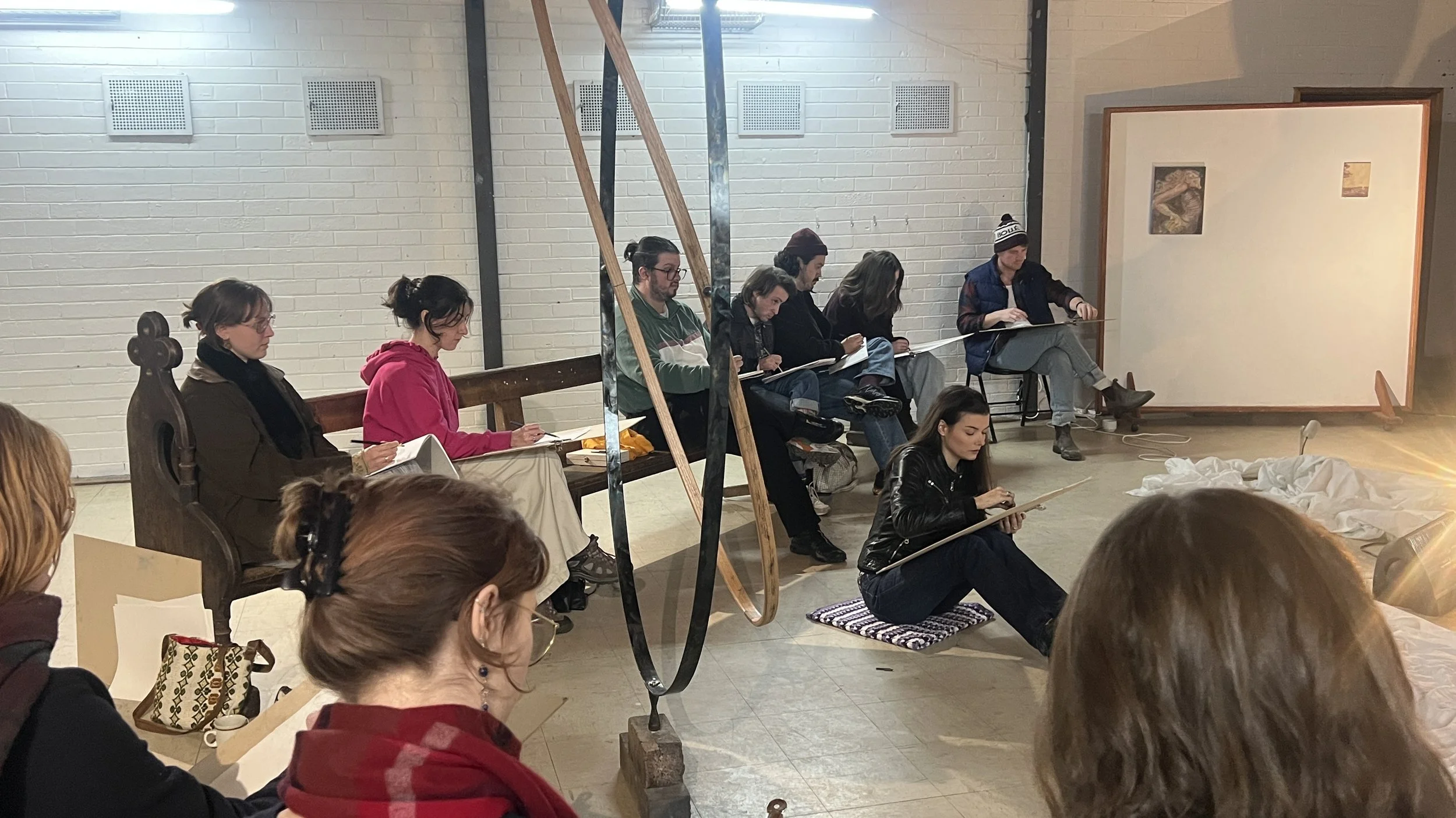People sitting in a classroom or workshop setting, some taking notes, others drawing or writing, with artwork and fabric on the floor and a large whiteboard in the background.