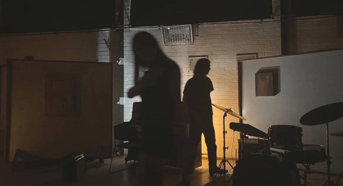 Silhouettes of two people in a dimly lit room with musical instruments, including drums and a keyboard, and artwork on the walls. for Oddany music Night