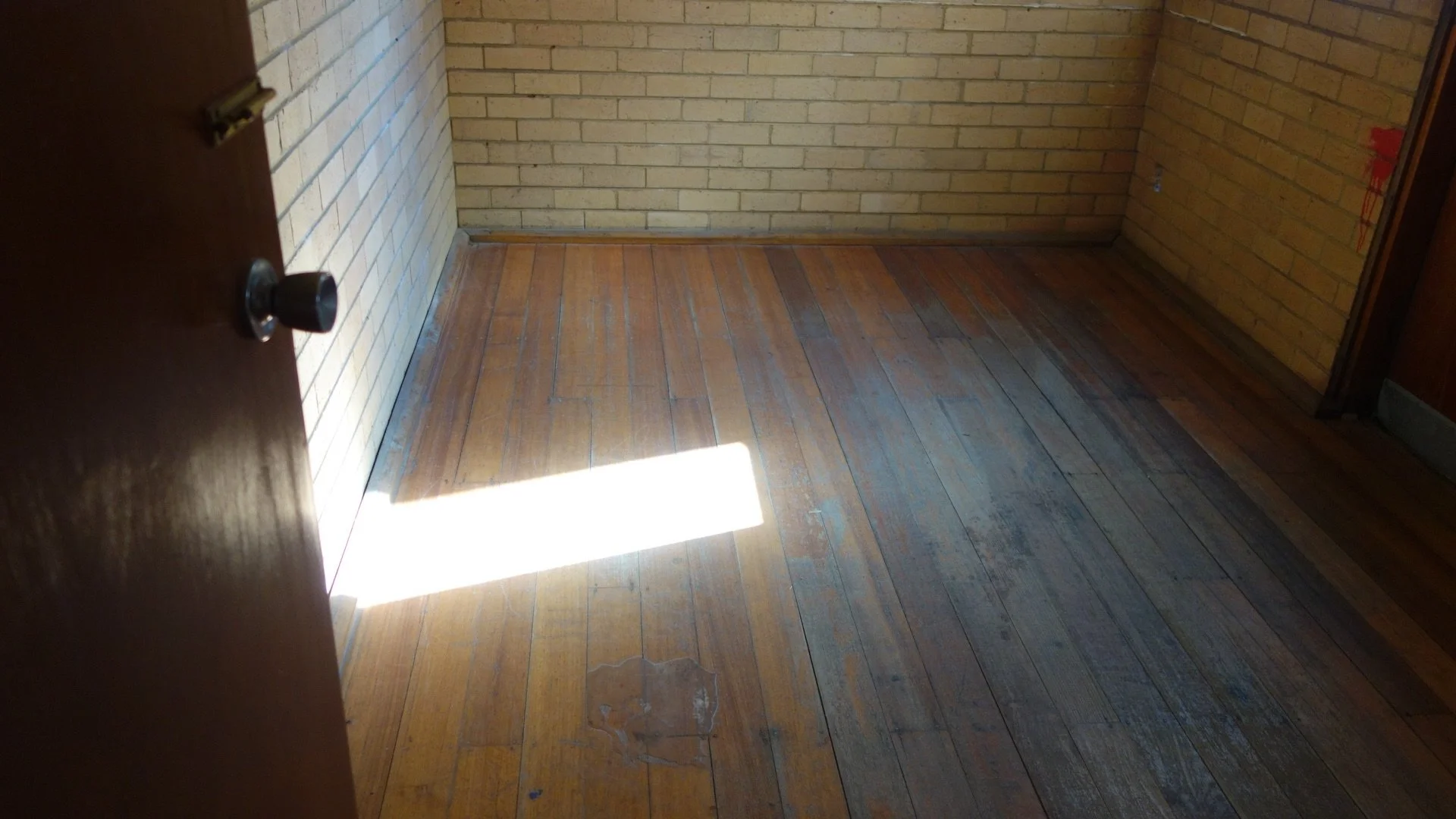 Empty room with wooden floor, brick walls, and sunlight entering through a window or door, casting a bright rectangular patch of light on the floor.