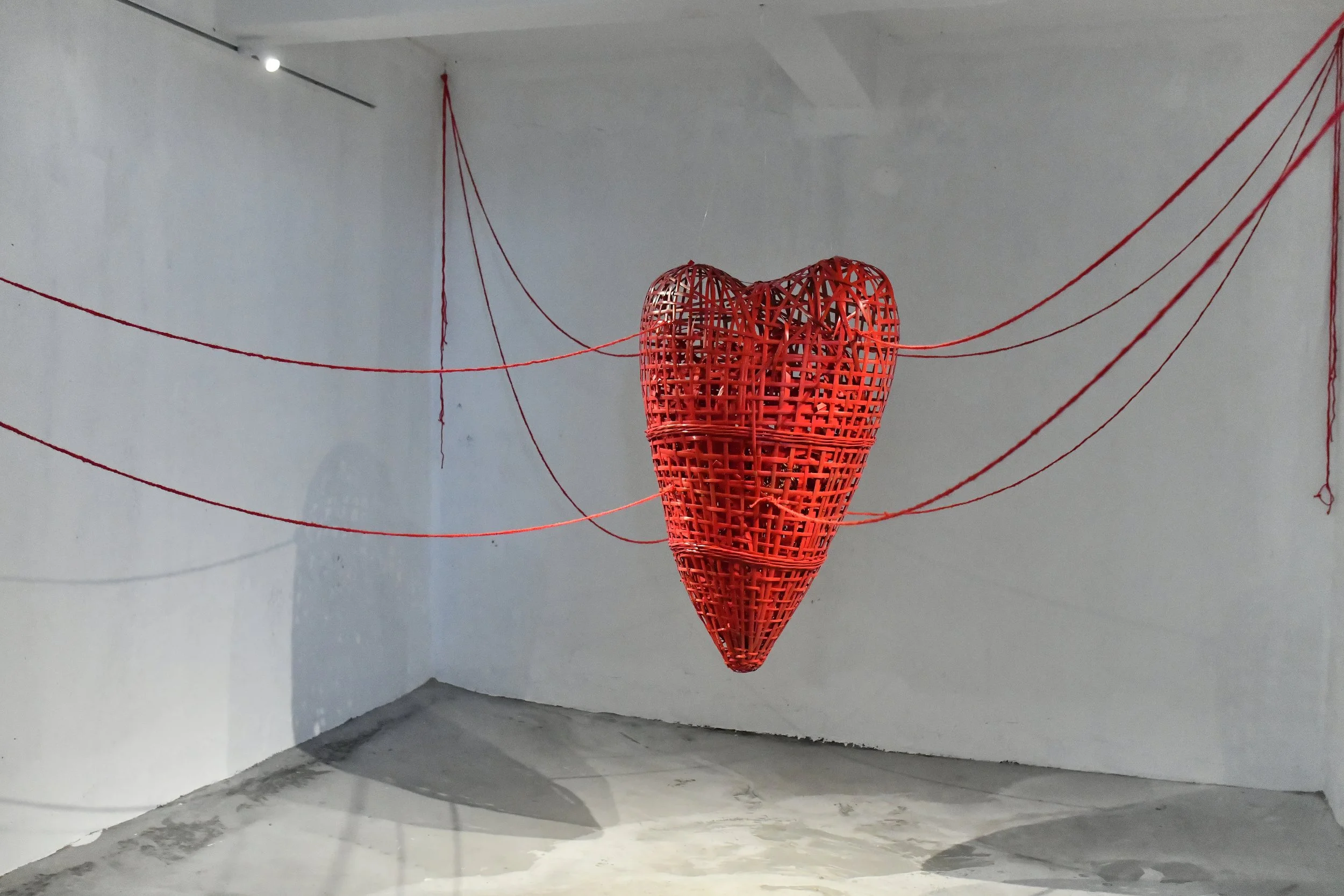 An art installation featuring a large, red, woven heart suspended in the center of a white room with red strings radiating outward.