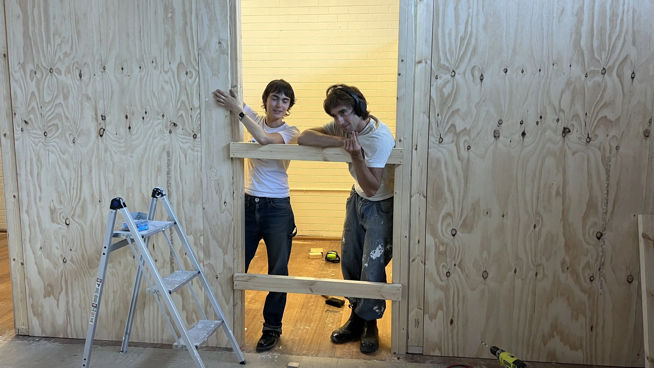 Two young men build a wall out of plywood. One is wearing headphones and shows a peace sign, while the other leans on the wall with one arm. There is a step ladder and tools nearby.