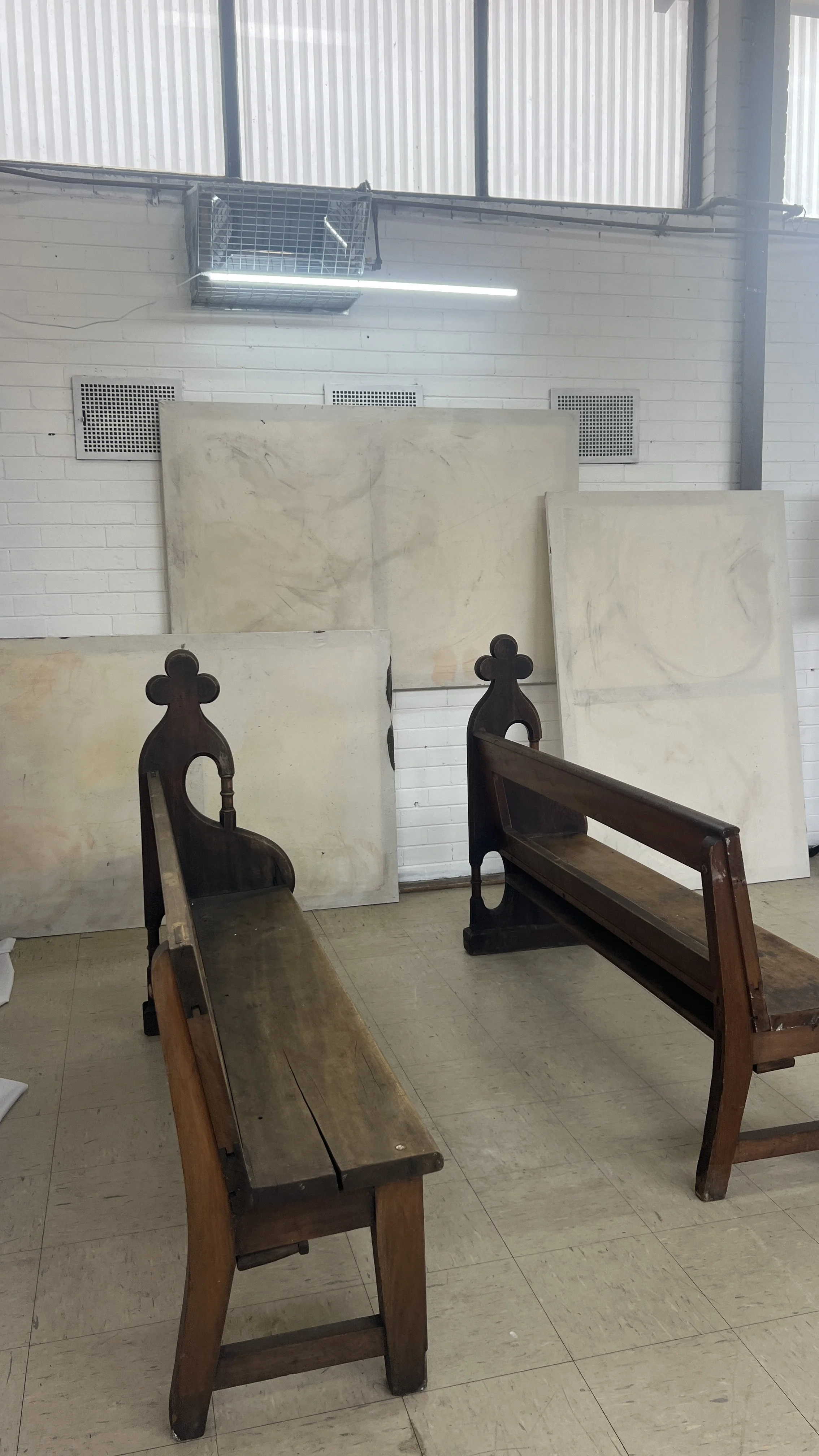 Two wooden church pews facing a set of large blank canvas paintings in an art studio with white brick walls.