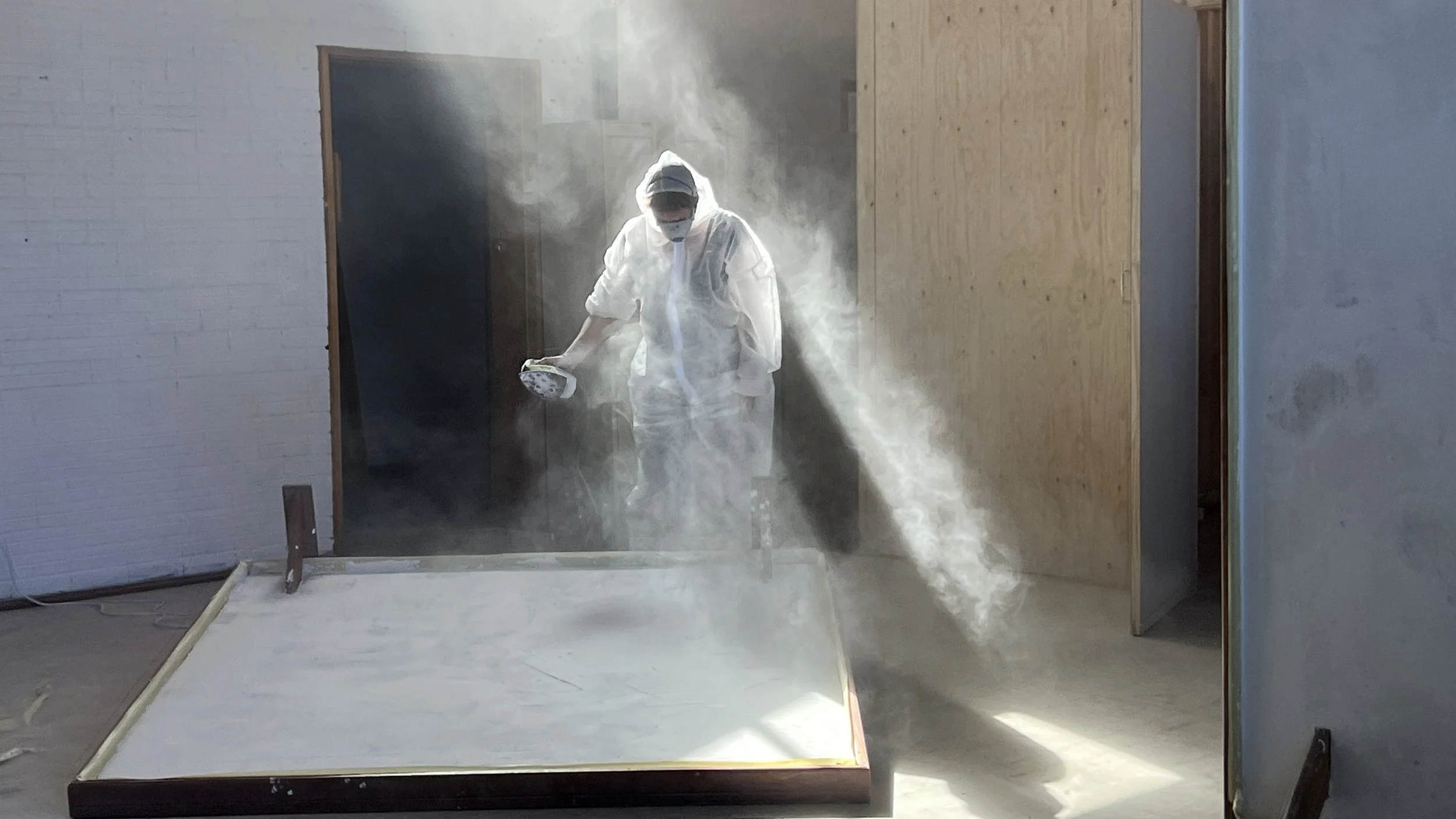 A person dressed in white protective suit, mask, and hood, spraying white powder or dust on a large tray in a room with plain white and wooden walls, amidst a cloud of dust or powder.
