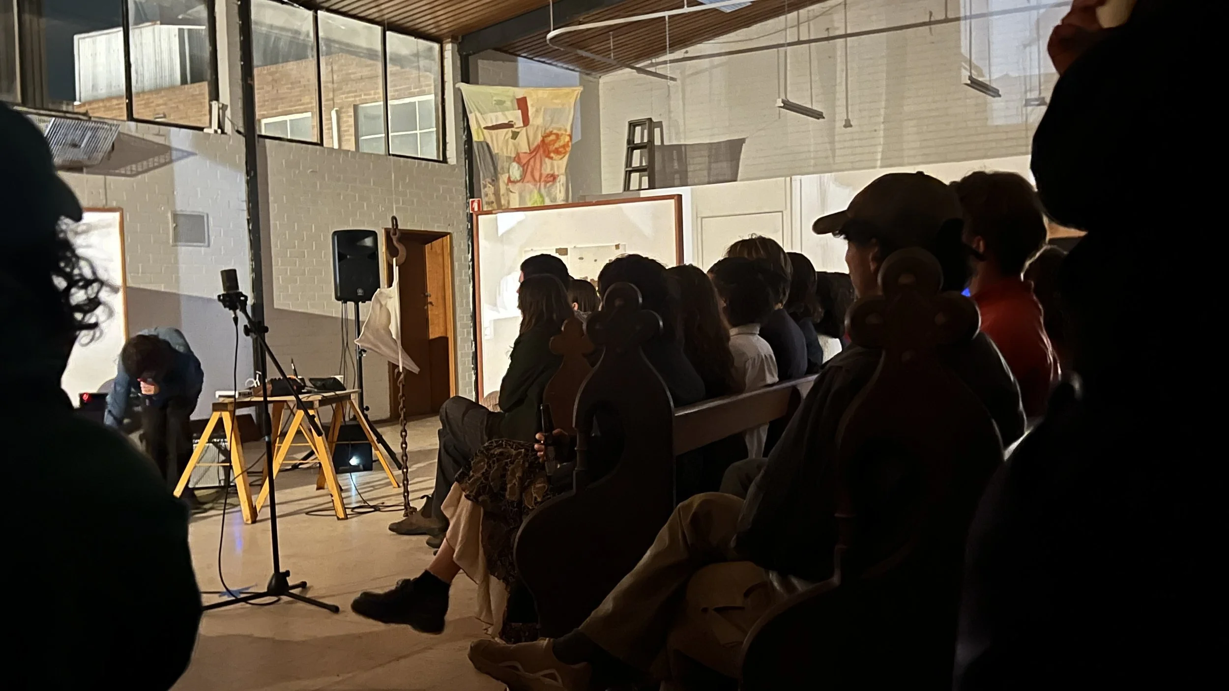 People seated in a dimly lit room watching a presentation or performance, with some equipment and a person setting up at the front of the room.
