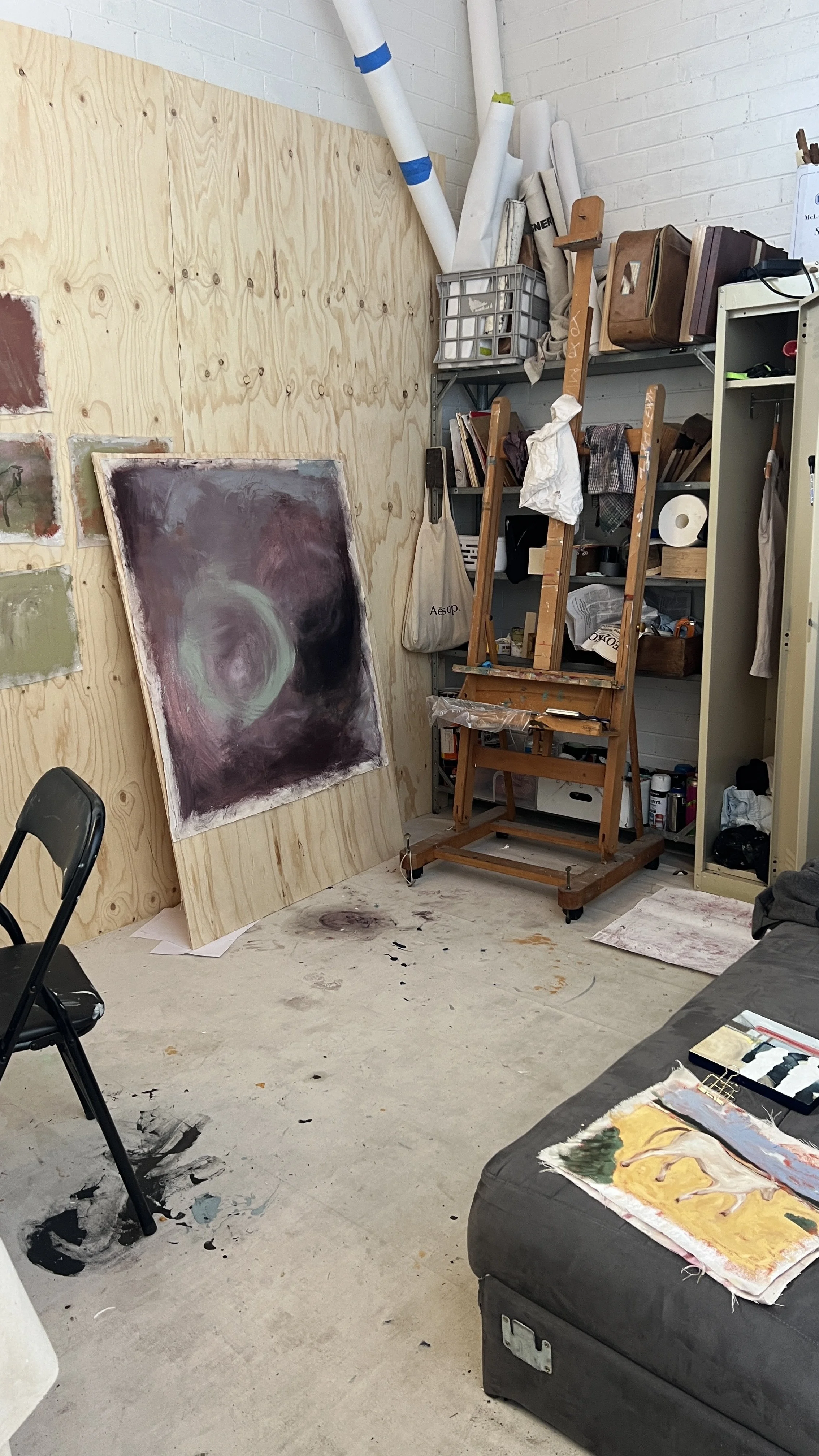 Art studio with a large abstract painting leaning against a wooden wall, an easel, and various art supplies on shelves and the floor.