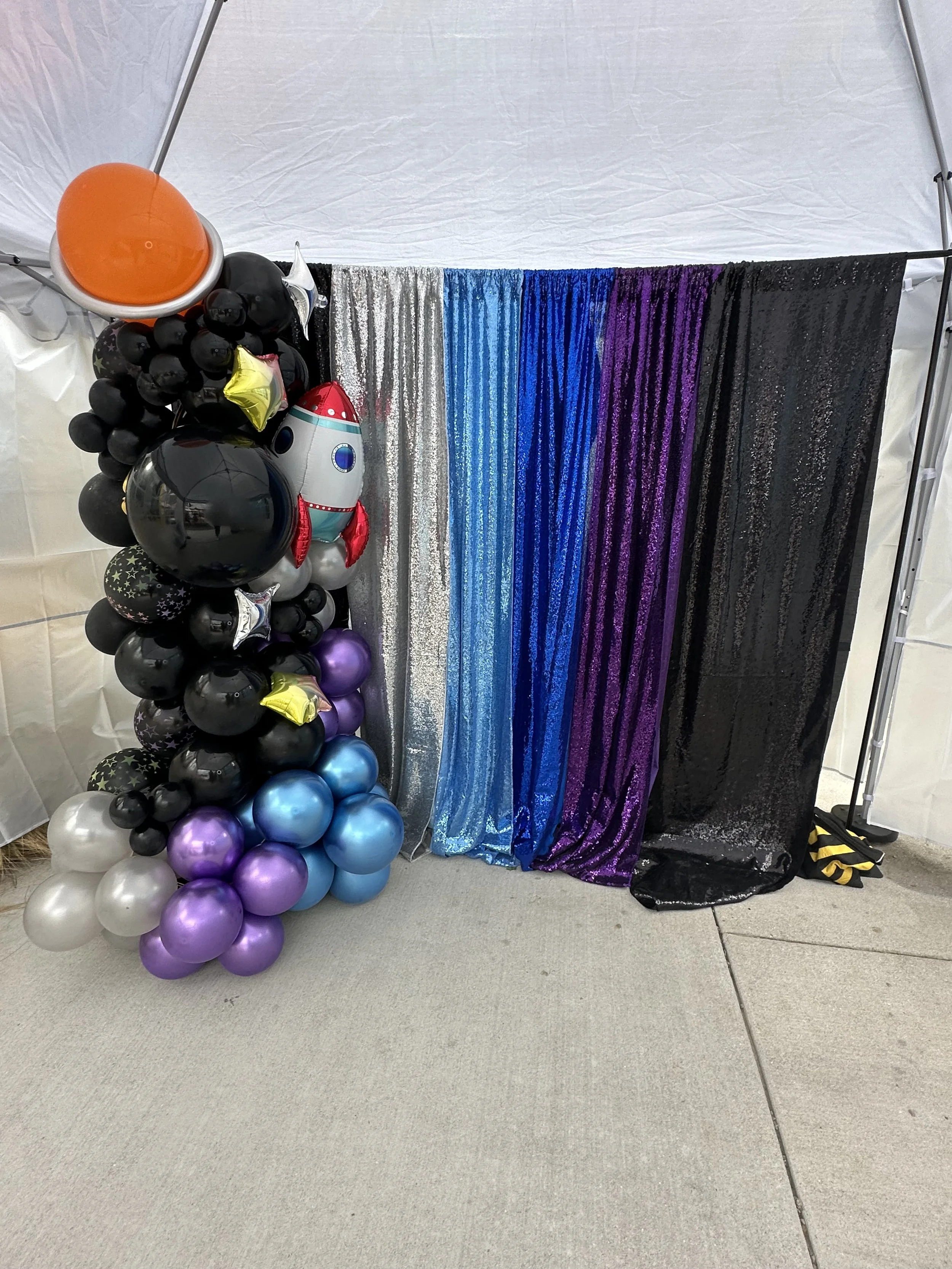 Wide angle shot of Honyes Booth Galatic Photo Booth rental set up in Hampton Roads Virginia with black, White/opal, blue & purple galactic/outerspace balloon arch with stars, a rocket ship & planet balloons. Outdoor set up with tent. 