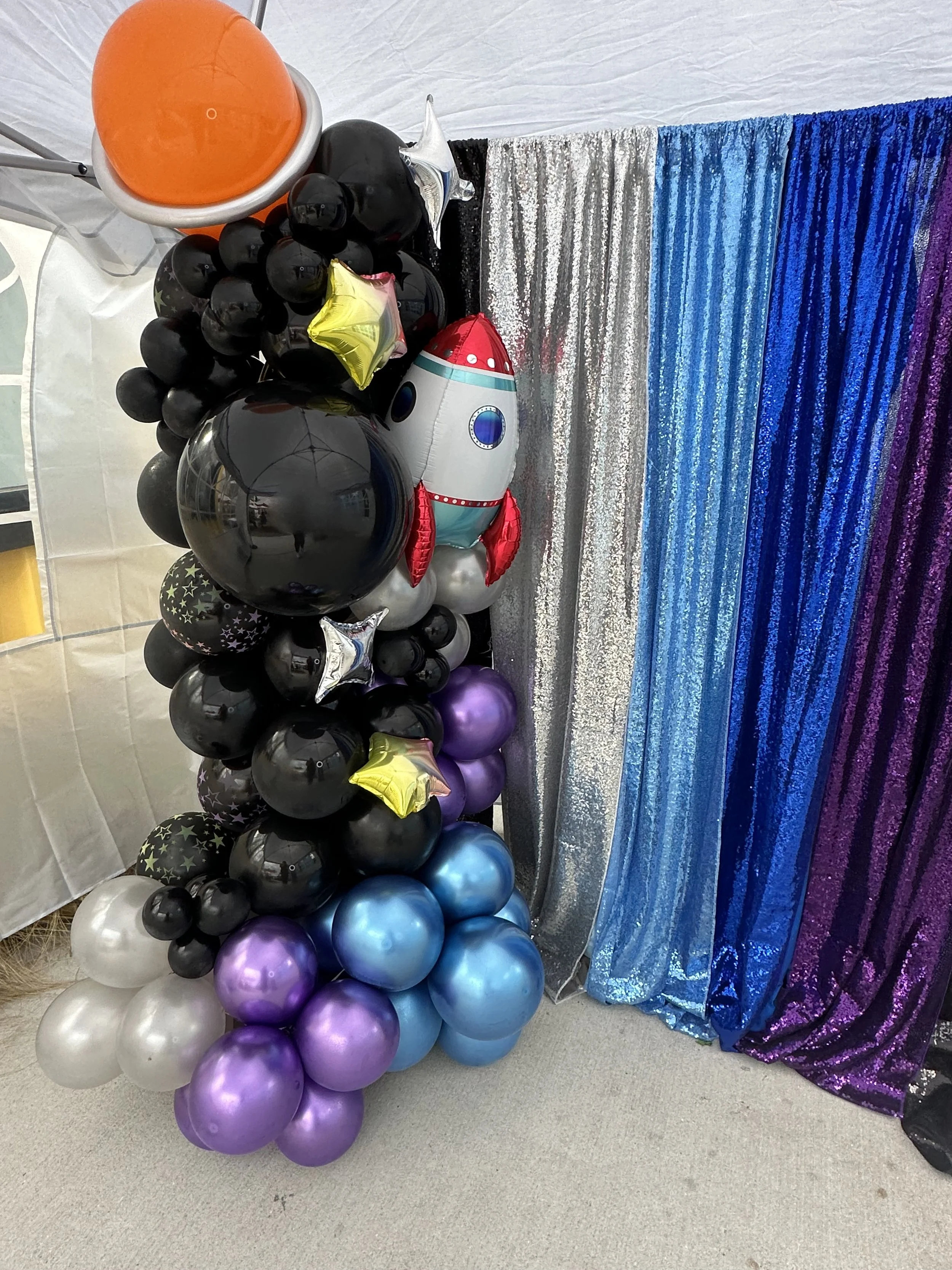 Honyes Booth Galatic Photo Booth rental set up in Hampton Roads Virginia with black, White/opal, blue & purple galactic/outerspace balloon arch with stars, a rocket ship & planet balloons. 