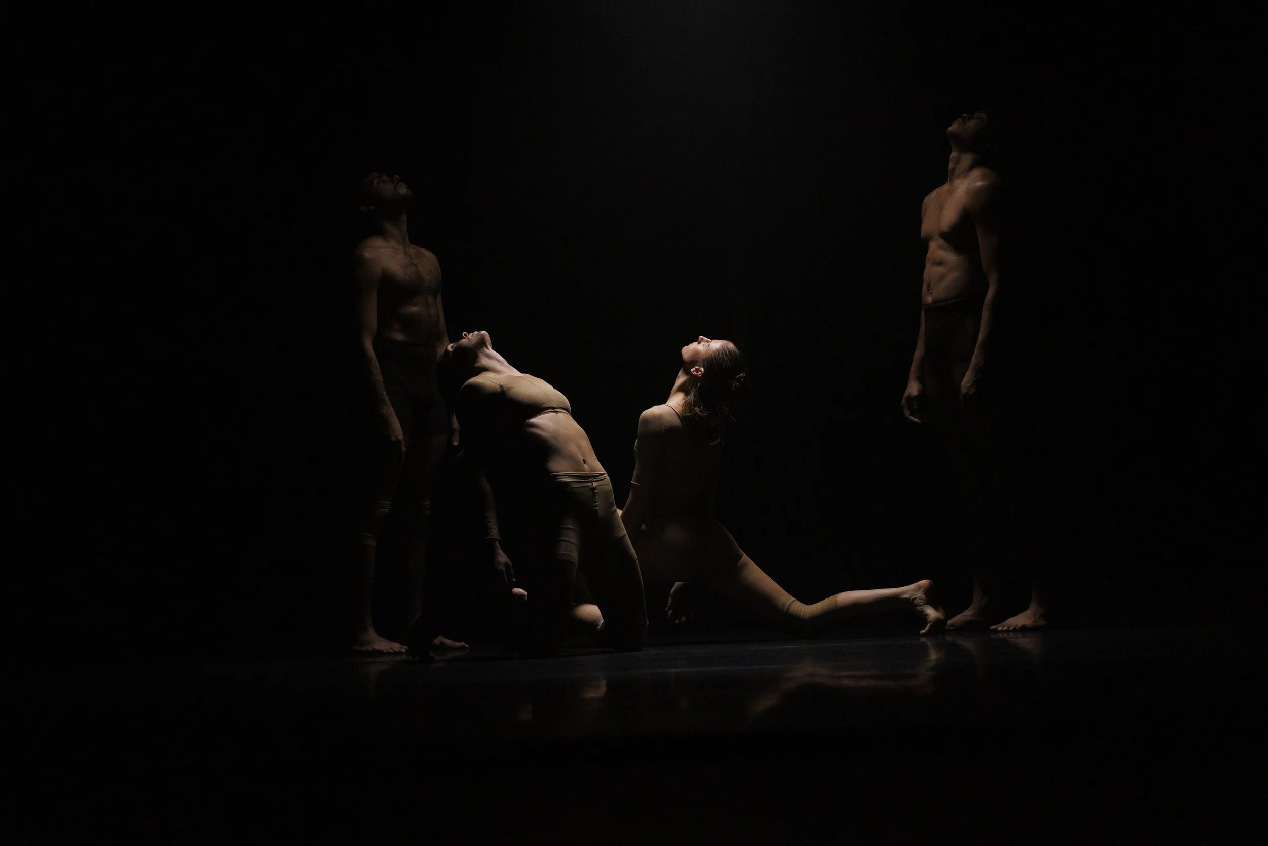 Five dancers in minimal light perform on dark stage, with three standing at the sides and two kneeling or stretching in the center.