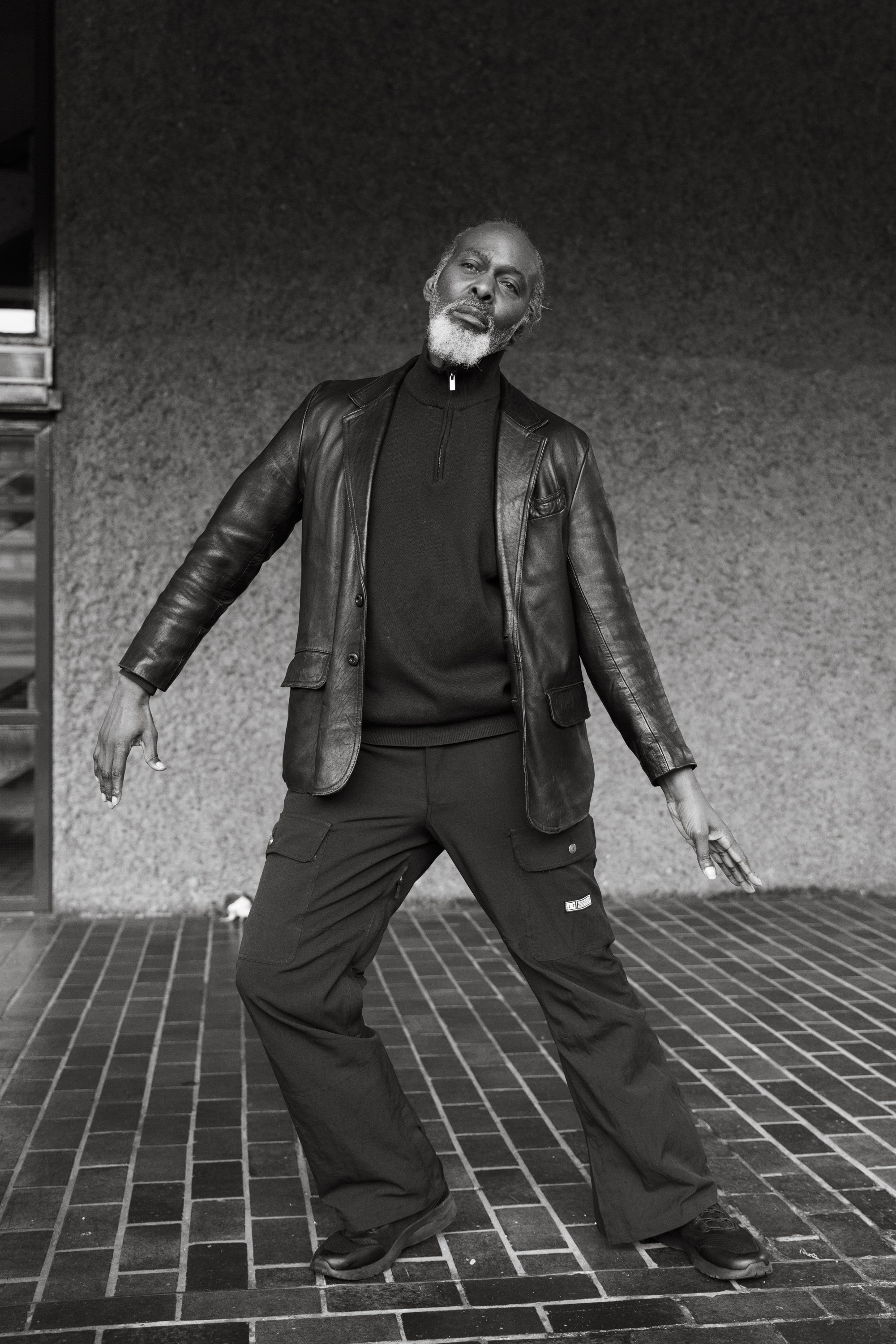 An older man with a beard standing outdoors on a brick surface, wearing a leather jacket, a black zip-up shirt, and black cargo pants, striking a pose with arms outstretched.