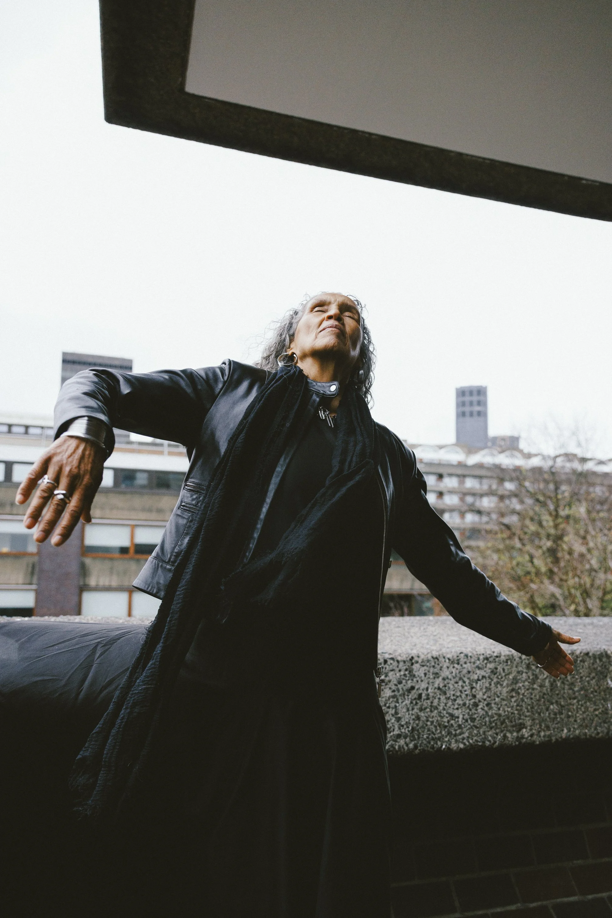 Woman with gray hair standing outdoors with arms outstretched, wearing a black leather jacket and scarf, looking upward with a serene expression, overcast sky, city buildings in the background.