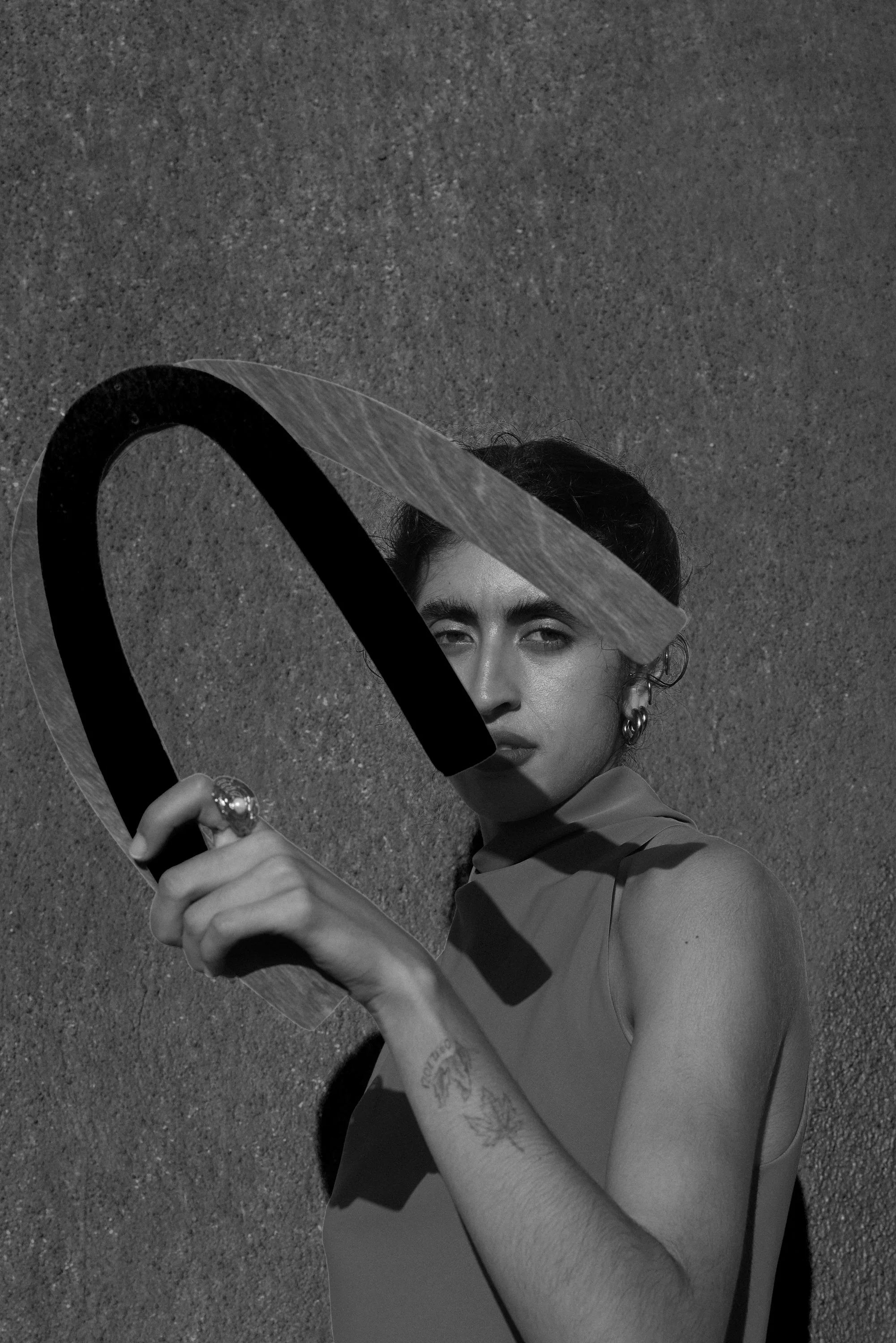 A black and white photo of a woman holding a large, stylized letter 'U' made from a carved material with a shadow cast on her face and shoulder. She has dark hair, earrings, and appears to be standing against a textured wall.