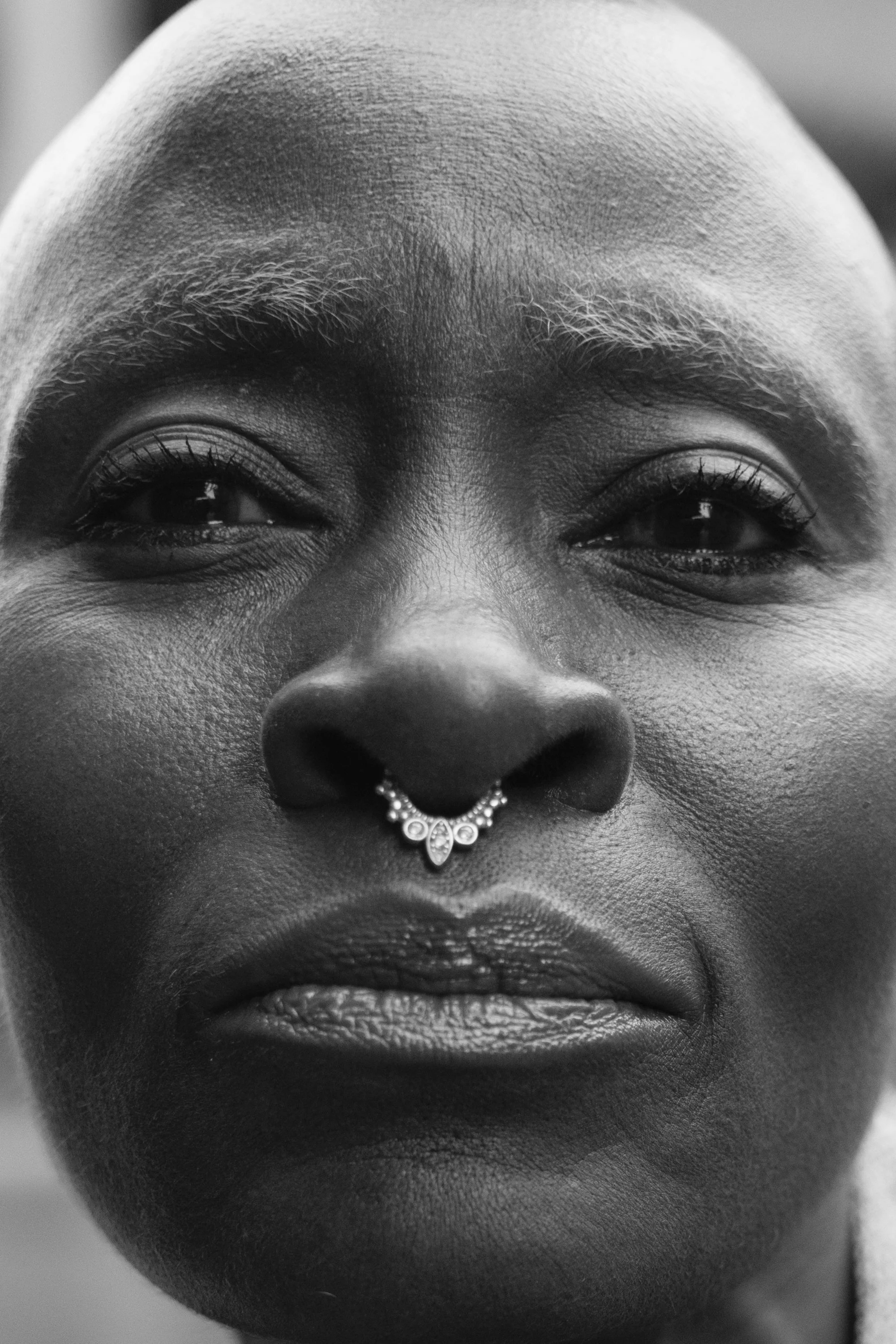 Close-up black and white photo of a person's face, highlighting the nose with a decorative septum piercing, full lips, and expressive eyes.