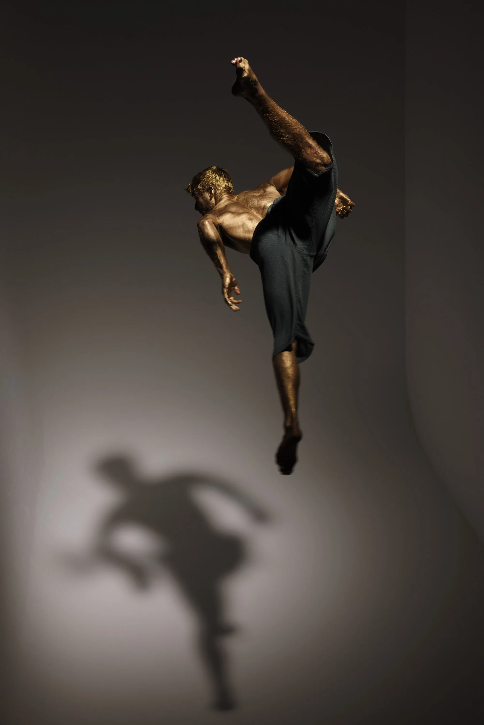 A shirtless man performing a high kick dance move, with his shadow cast on the wall behind him.