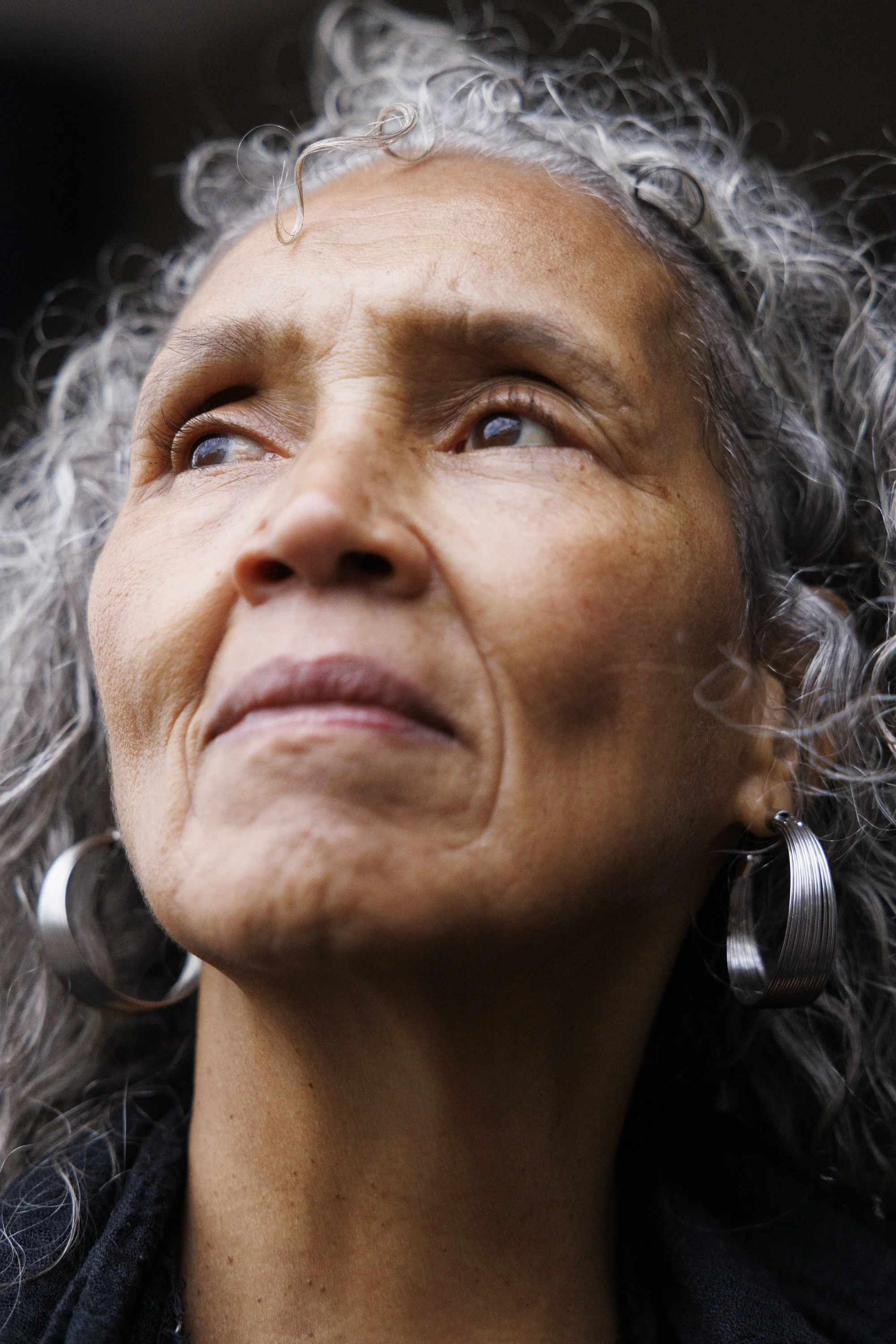 Close-up of an older woman with curly gray hair, looking upwards. She has hoop earrings and a subtle expression.