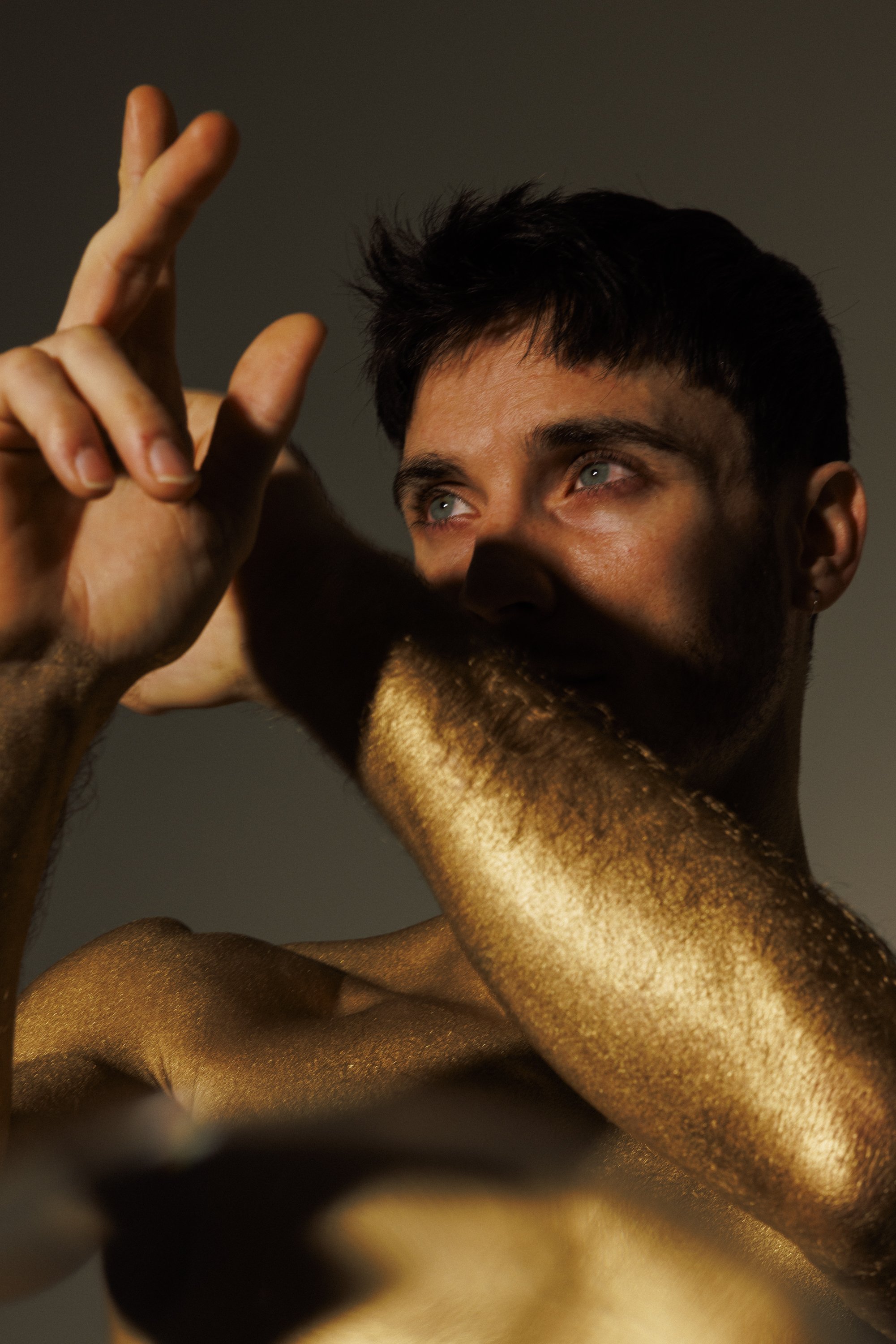 A young man with dark hair and blue eyes has his face and arm painted with gold metallic paint, looking upwards with a peaceful expression.